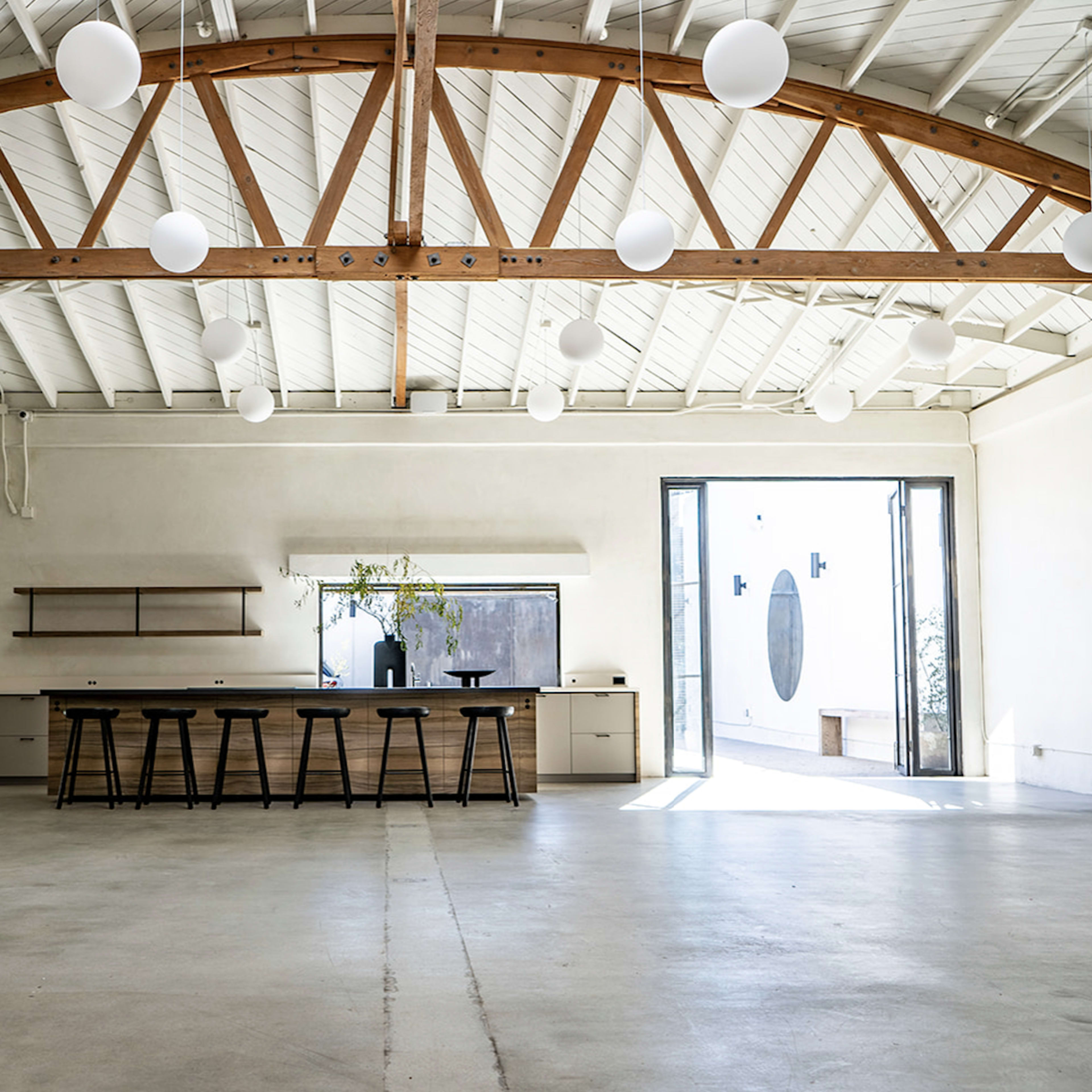 A spacious, minimalist interior with concrete flooring, a wooden beam ceiling, and a long counter with stools.