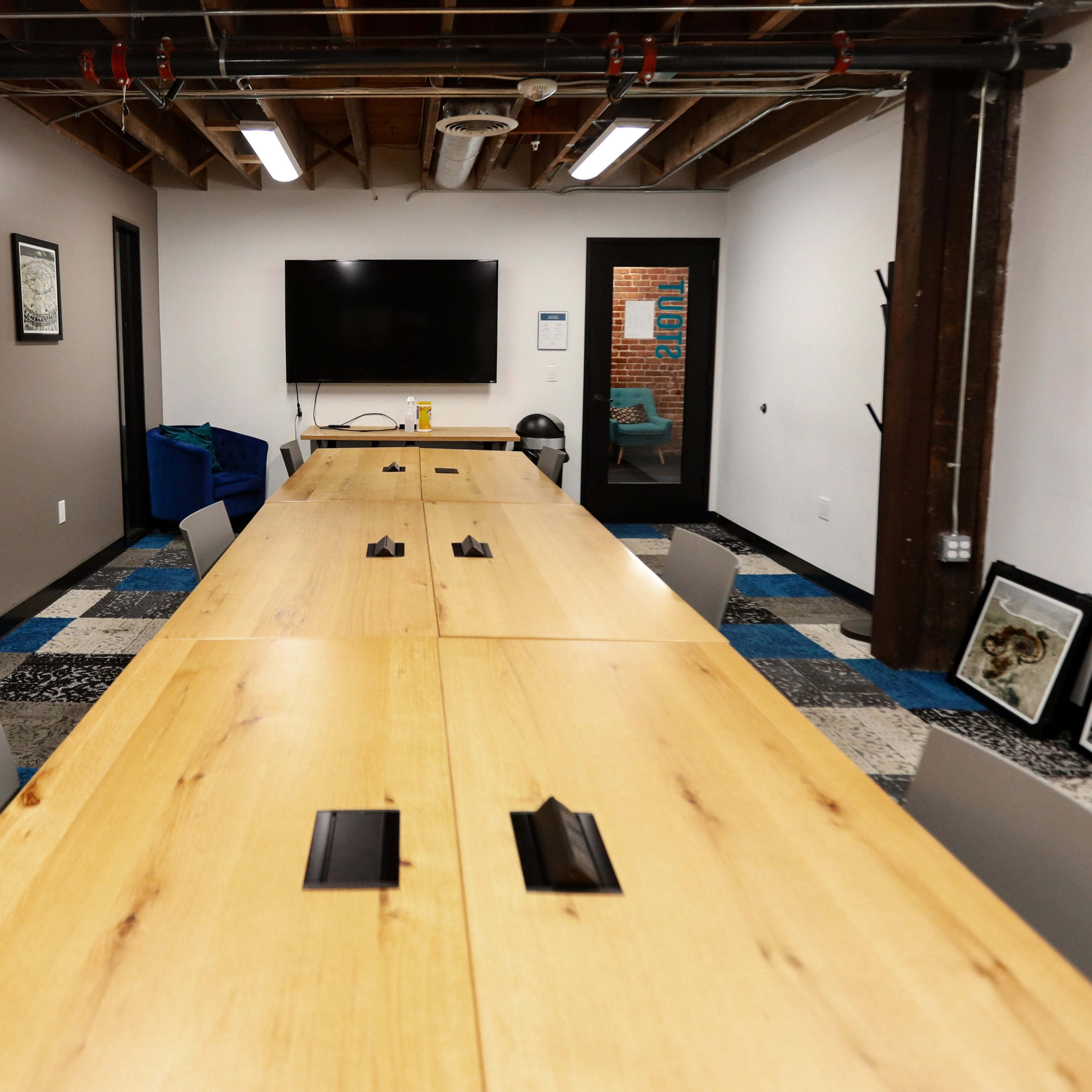 A modern conference room with a long wooden table, chairs, a large screen on the wall, and artwork framed on the walls.