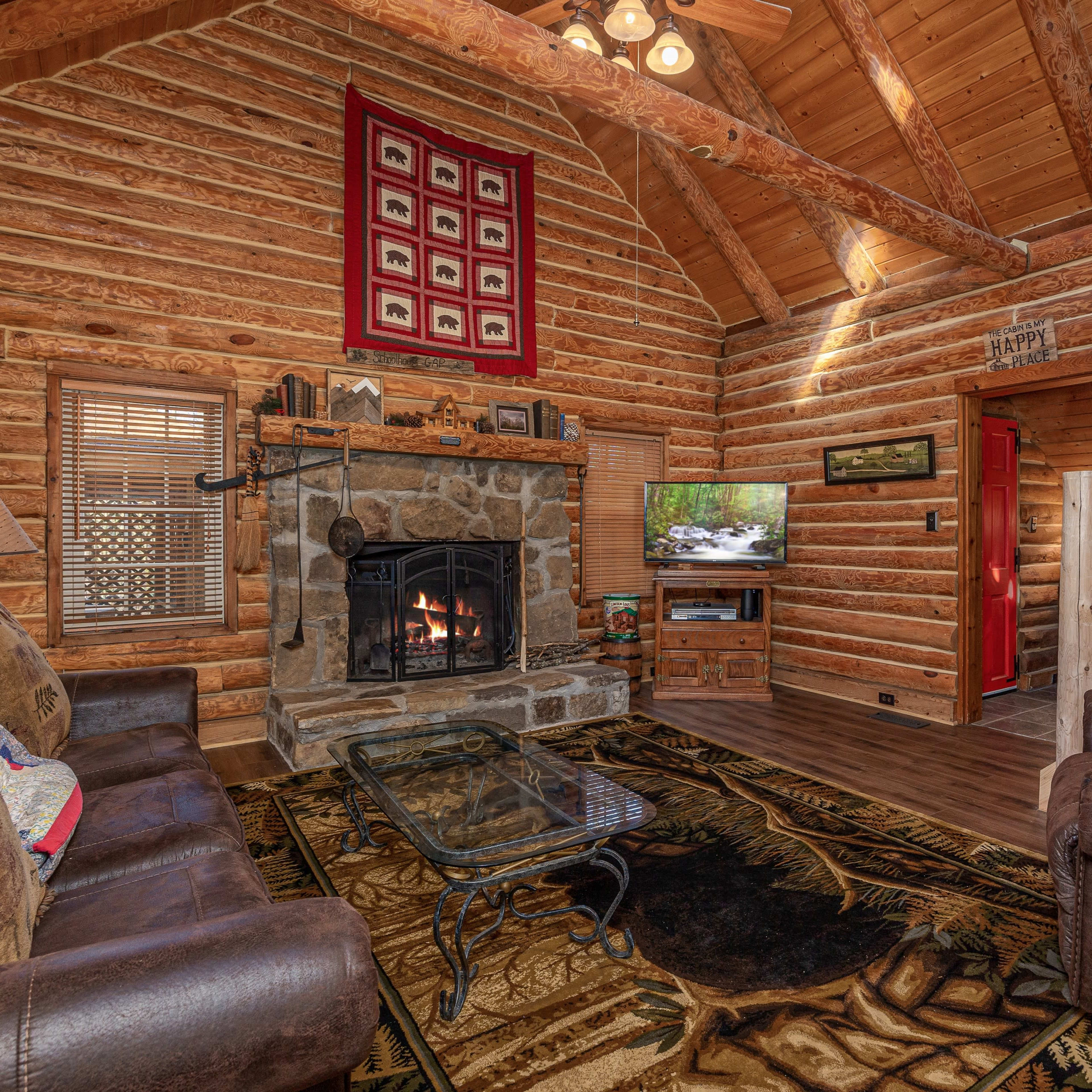 A rustic log cabin living room with a stone fireplace, wooden beams, and a staircase leading to a second level.