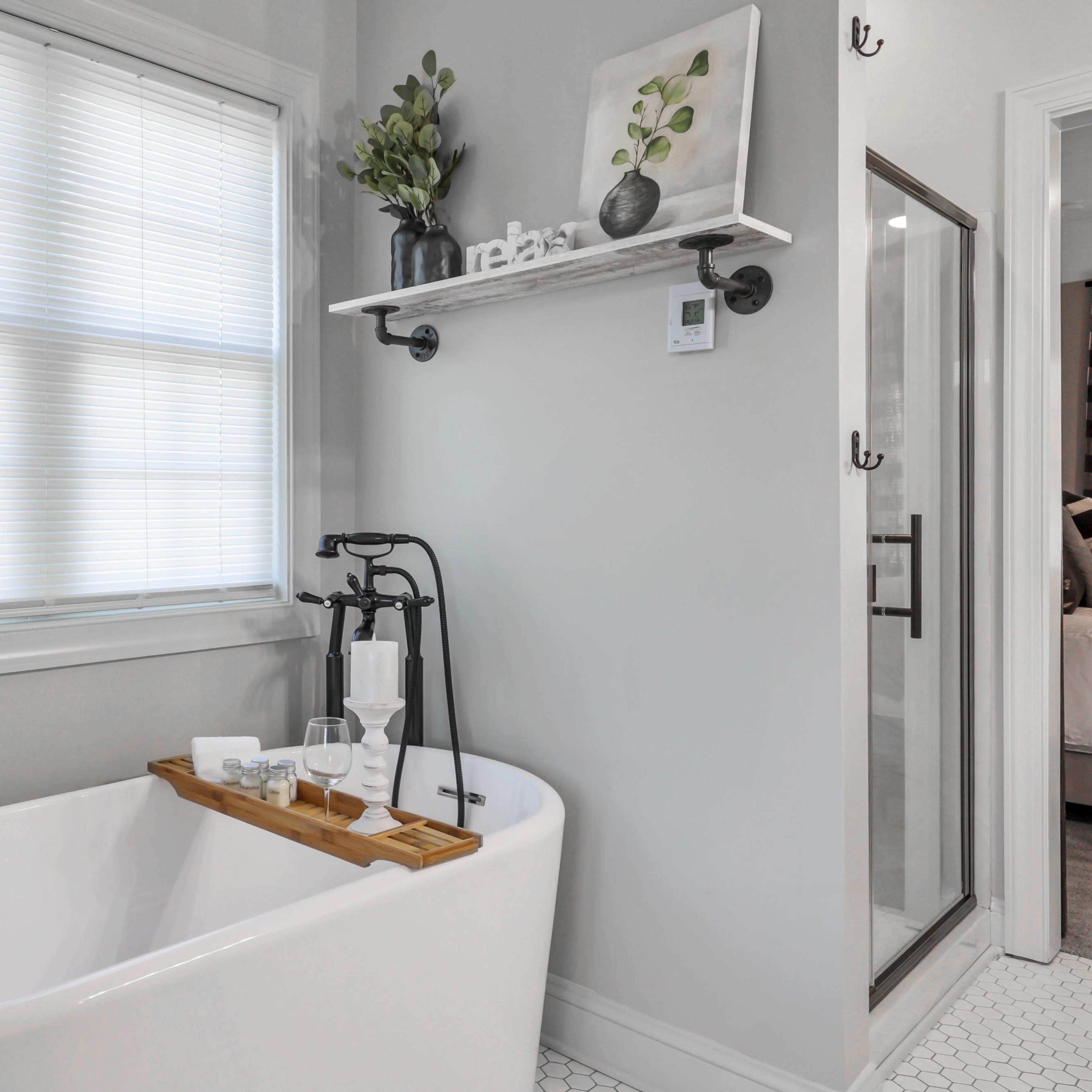 A freestanding white bathtub is positioned near a window with a wooden tray holding towels and glasses, complemented by a shelf with decorative items above.