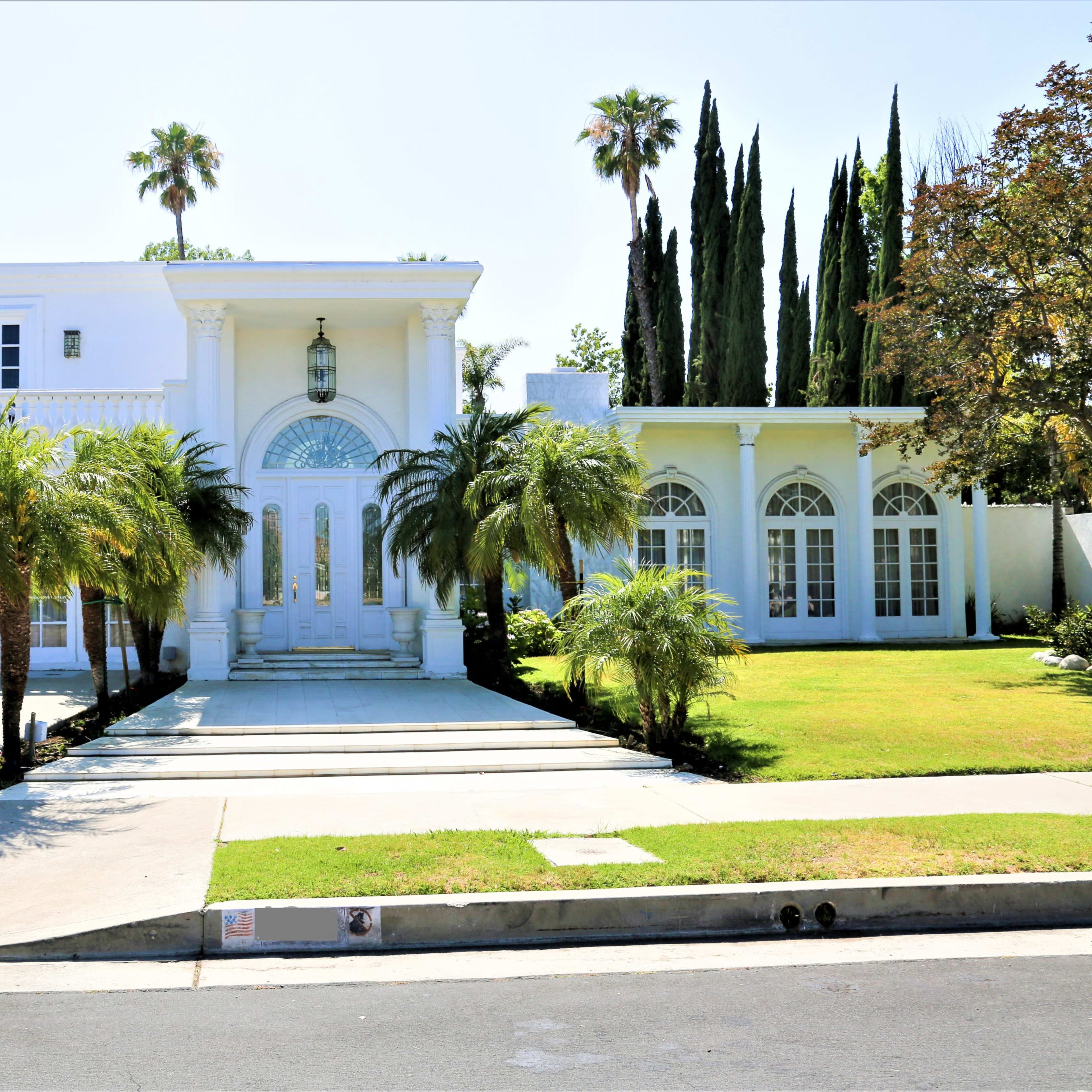 A large white house with a grand entrance, palm trees, and a well-maintained lawn is set at the end of a driveway.