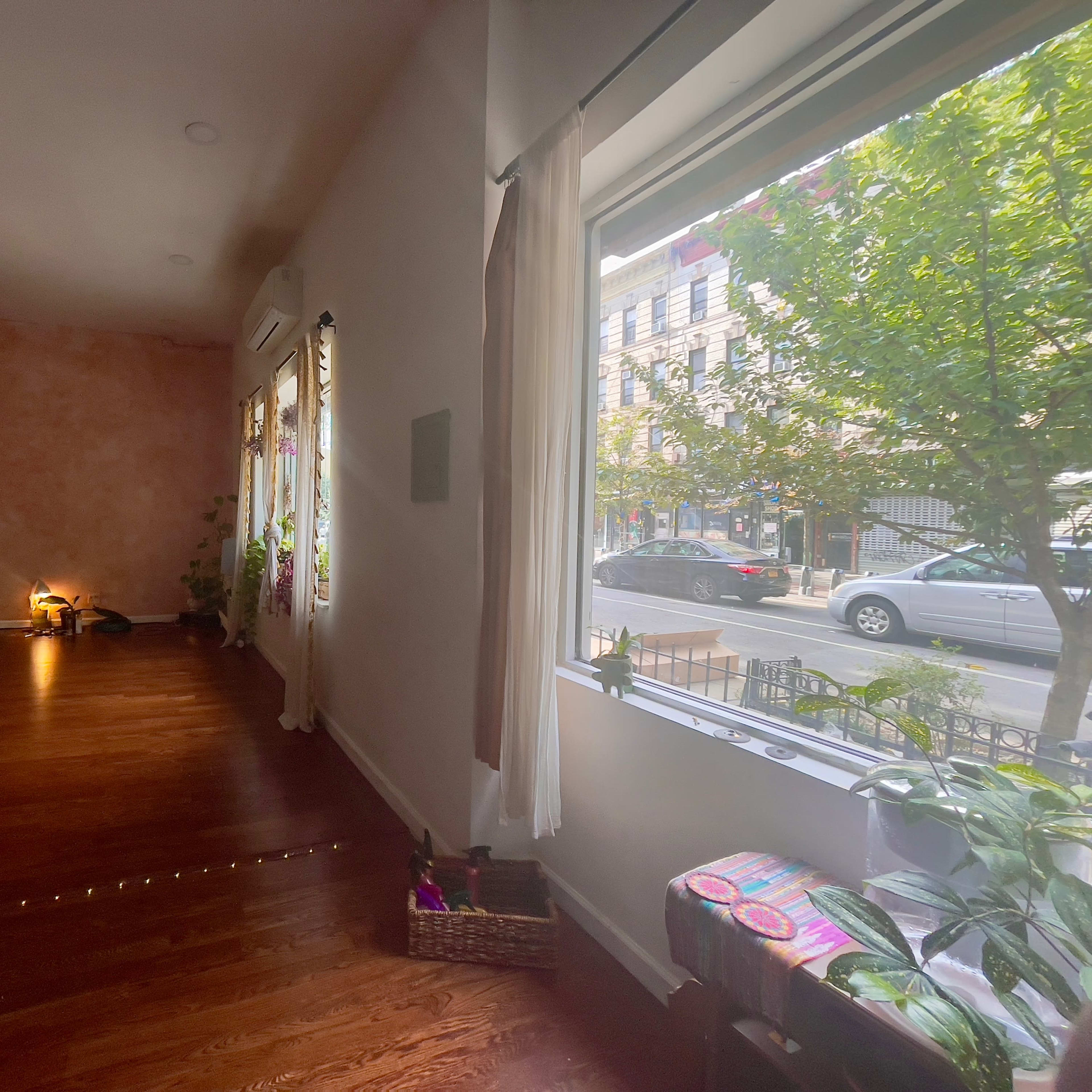 A spacious yoga studio with wooden floors, large windows allowing natural light, and a view of a street outside.