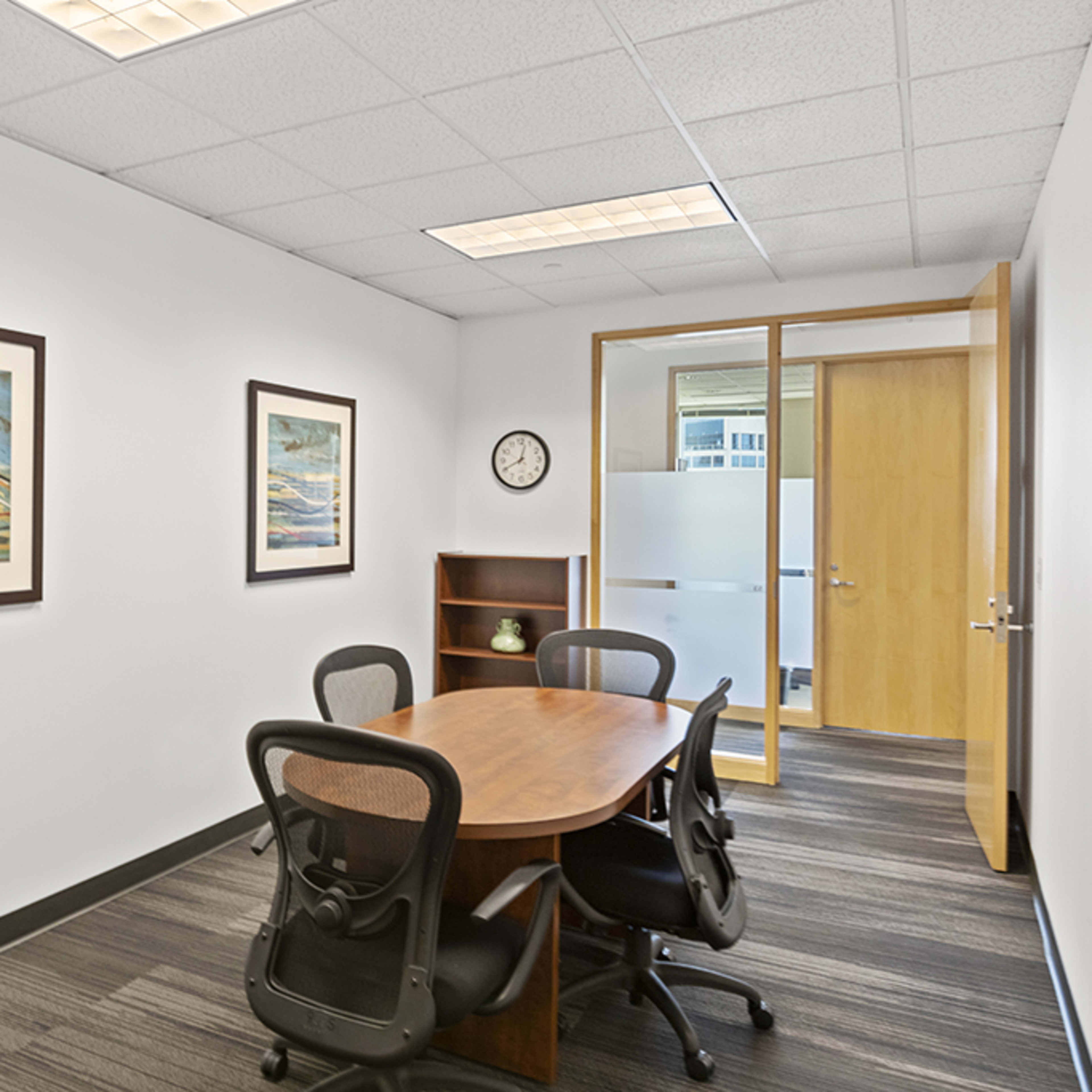 A conference room features a round wooden table surrounded by four black chairs, with two framed paintings on the white walls and a clock on the shelf.