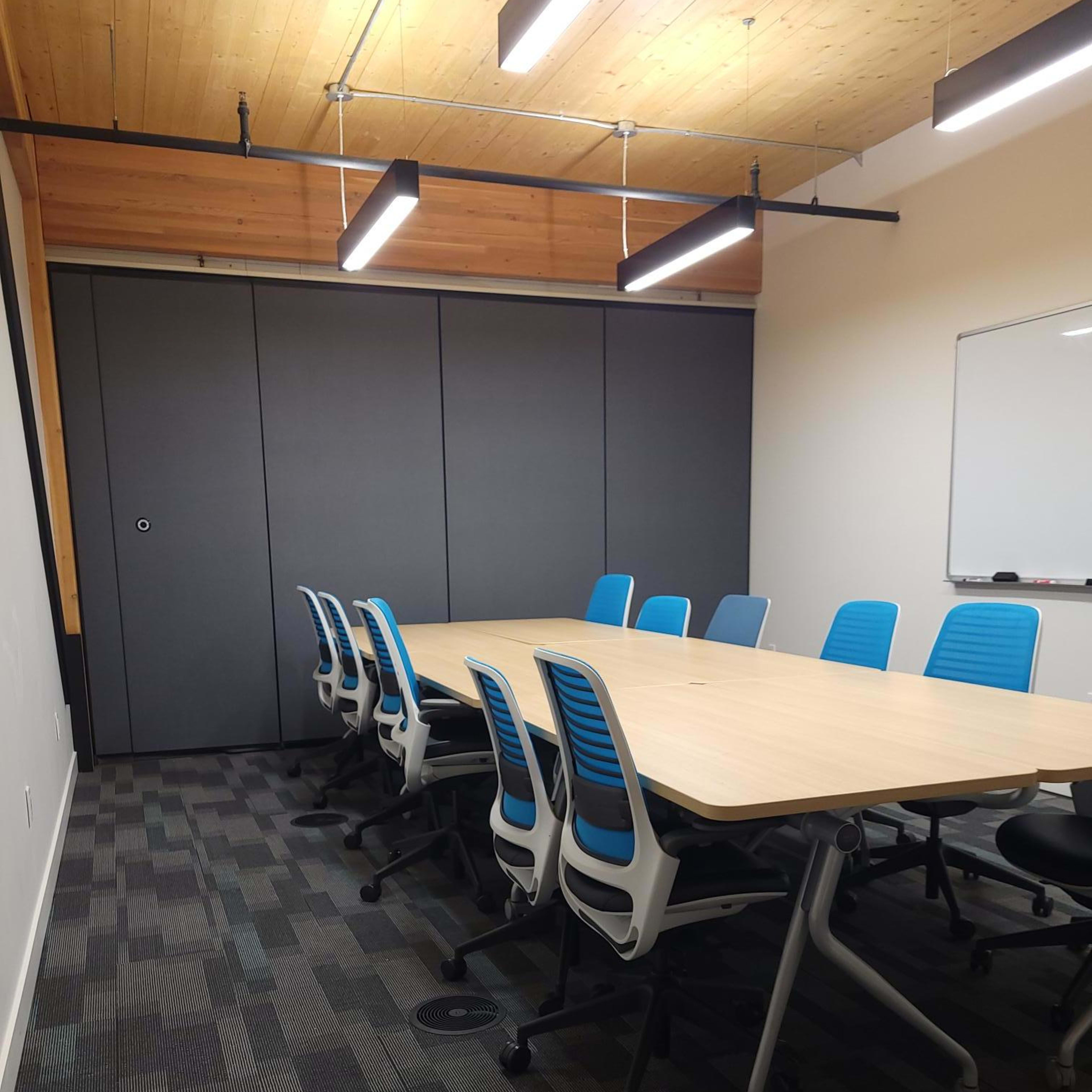 A modern conference room features a long wooden table surrounded by blue office chairs, with a whiteboard and storage cabinet in the background.