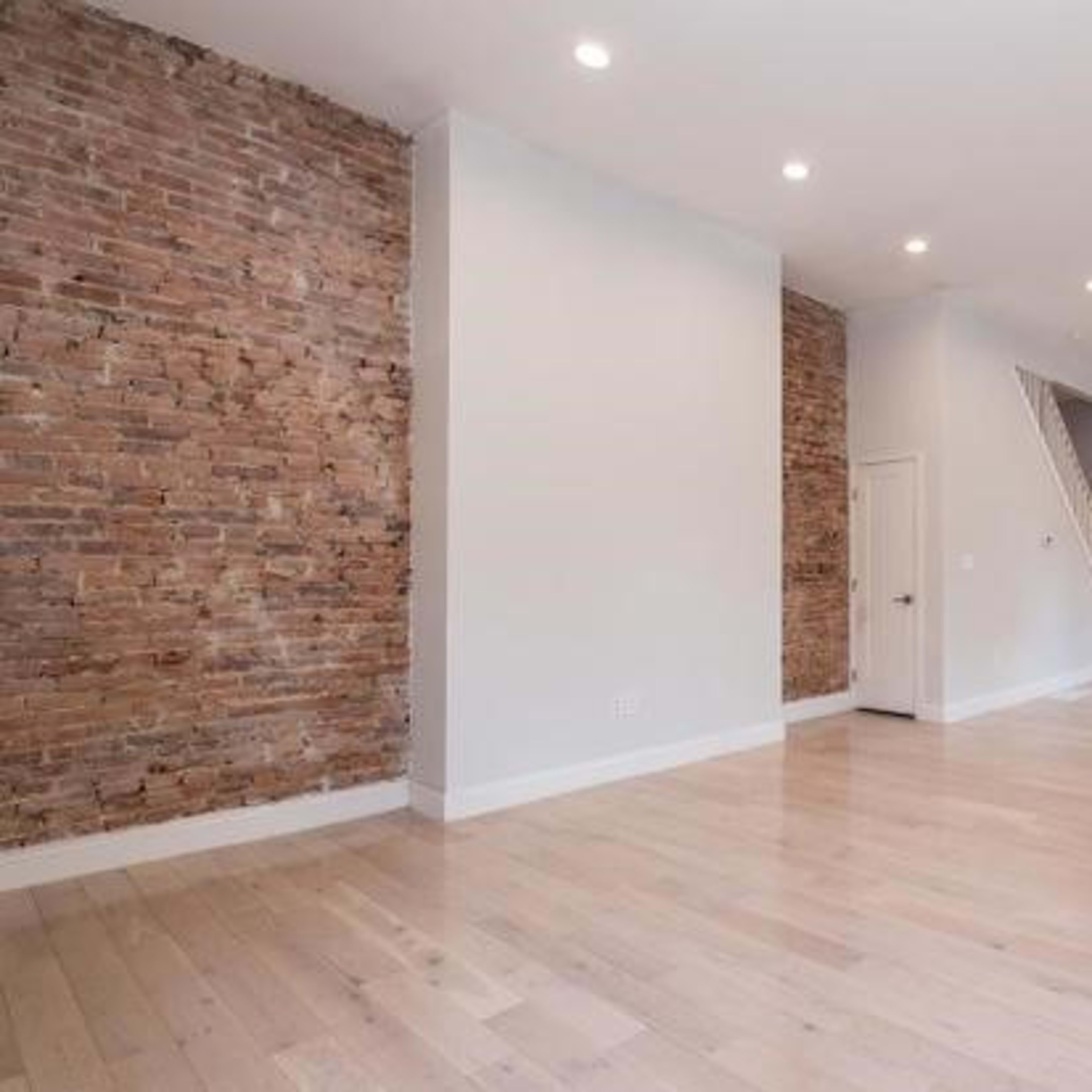 A spacious interior with a mix of exposed brick and smooth white walls, hardwood floors, and a kitchen area visible in the background.