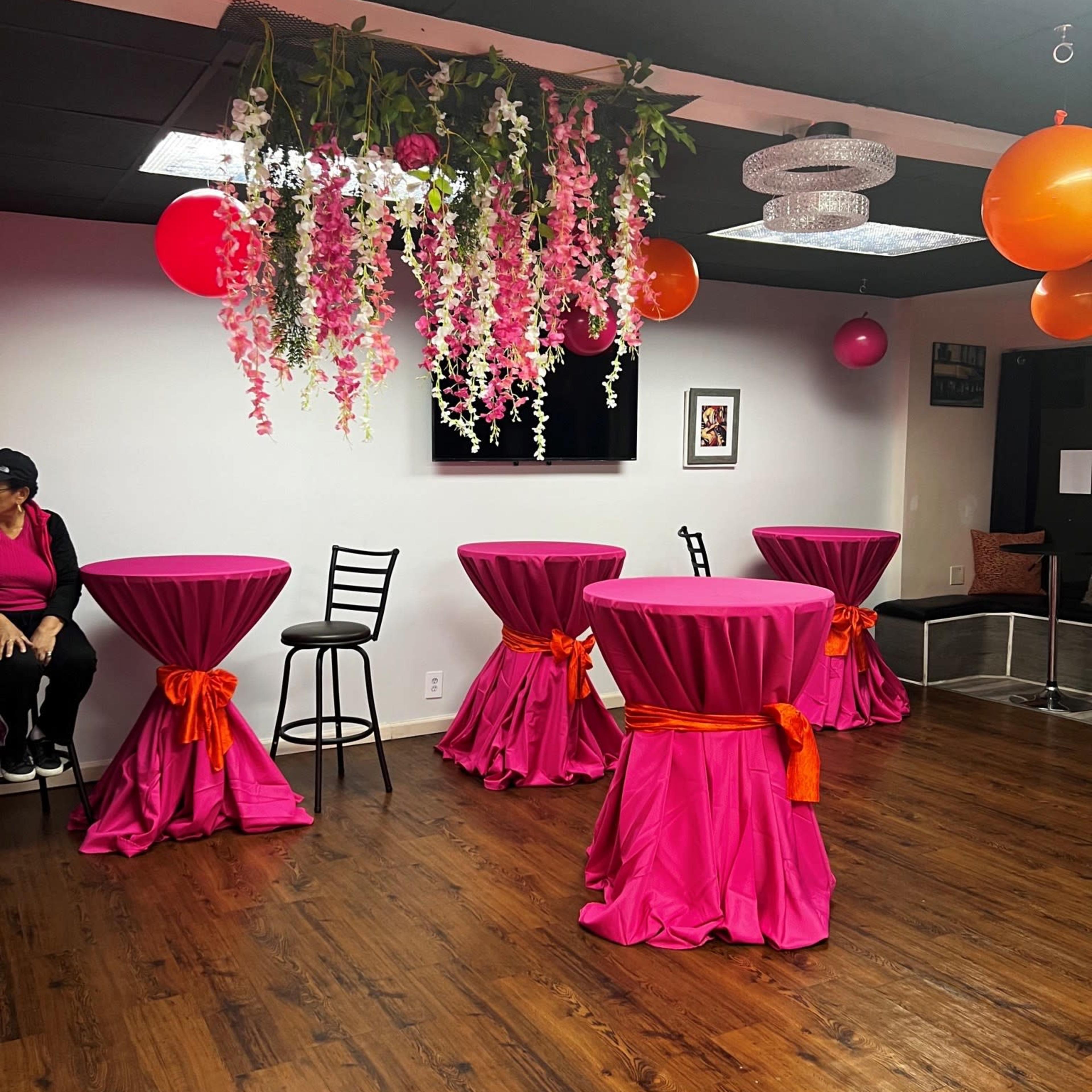 The interior features several round tables draped in pink tablecloths, adorned with orange sashes, amidst hanging decorations of flowers and balloons.