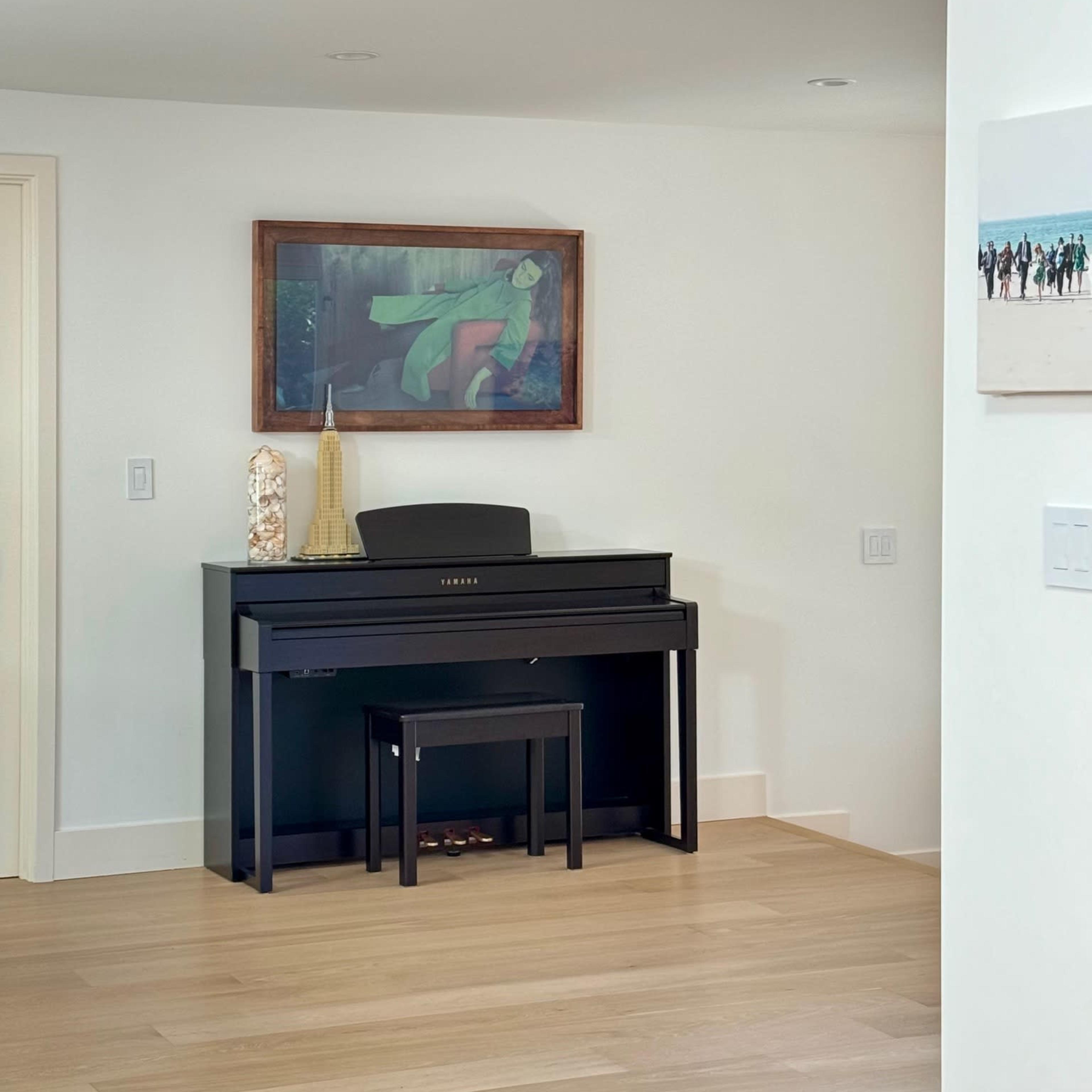 A digital piano with a matching bench is positioned against a wall, accompanied by a painting and decorative items on a nearby shelf.