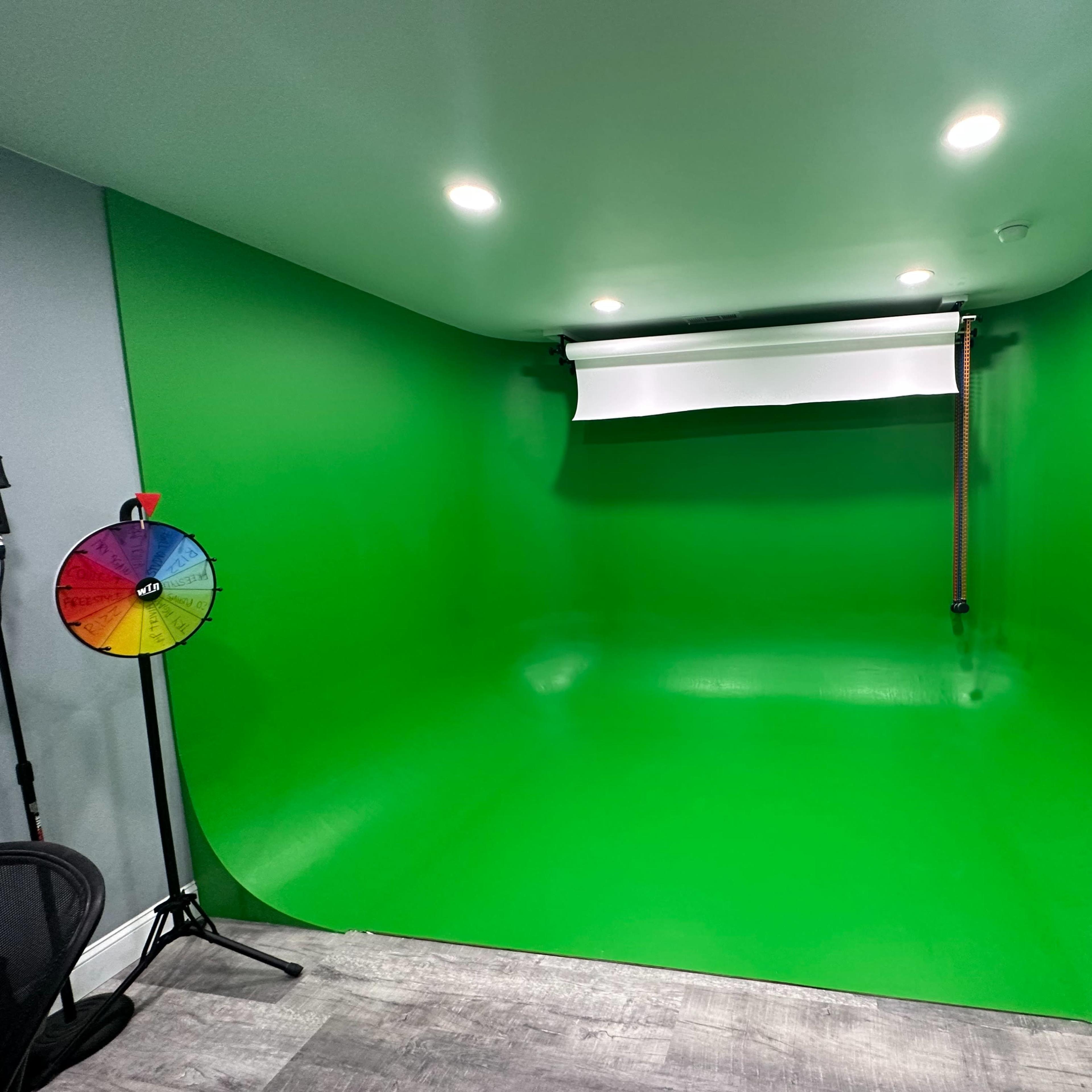 A green screen setup in a room, featuring a smooth green backdrop, overhead lighting, and a color wheel on a stand.