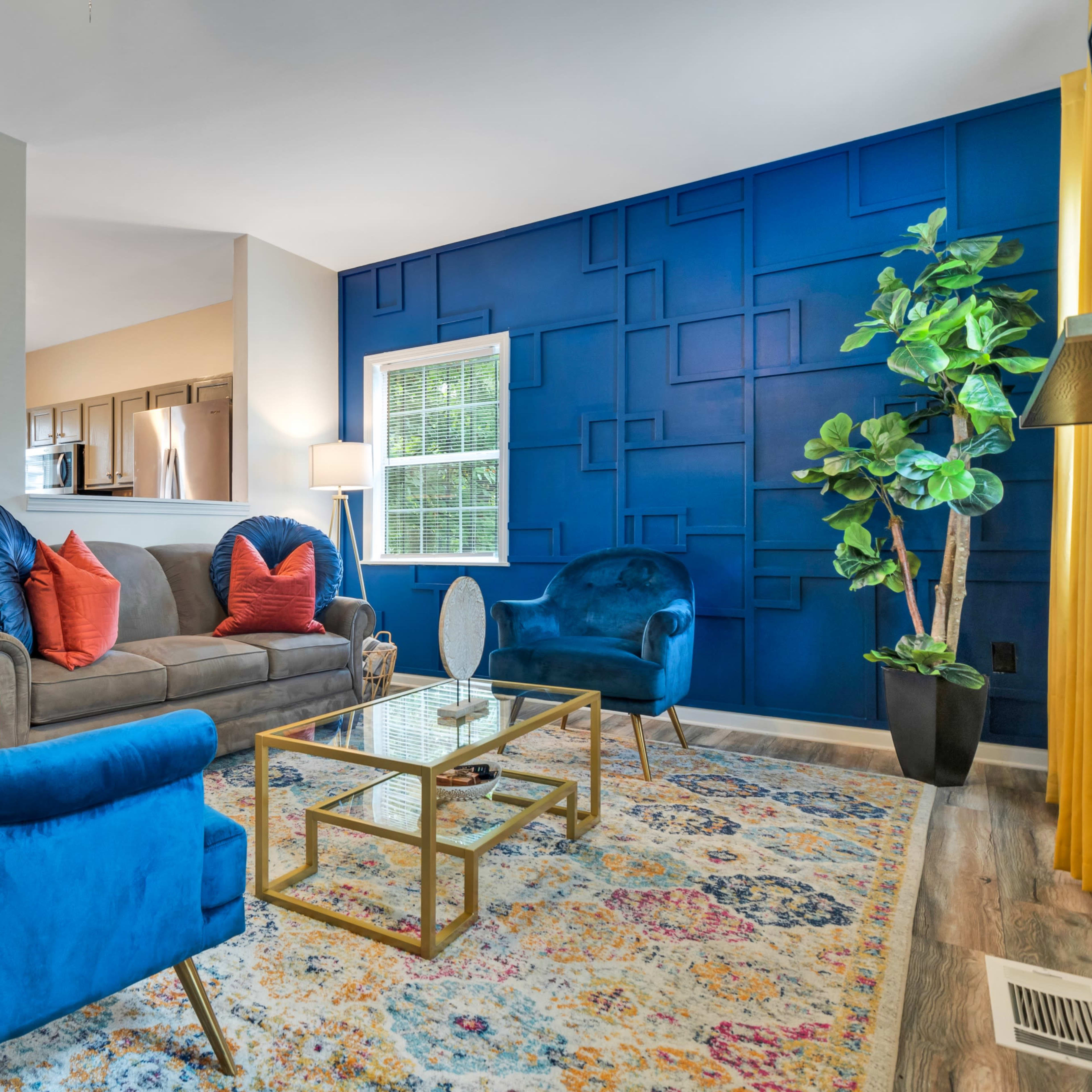 A living room featuring a blue accent wall, a gray sofa with orange pillows, two blue armchairs, and a glass coffee table, along with a plant and decorative artwork.