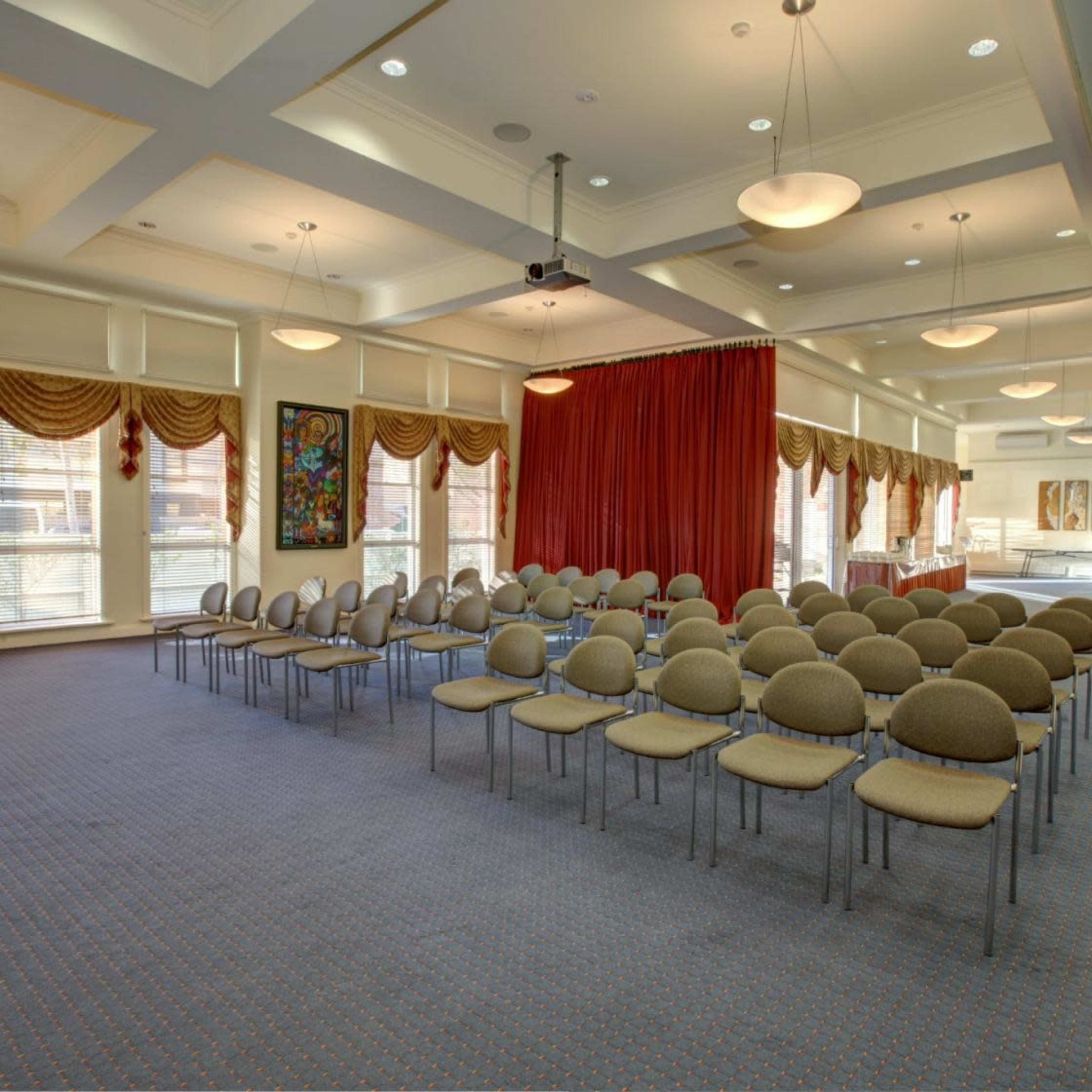 A meeting room set up with rows of circular chairs facing a red curtain, with large windows and decorative drapes along the walls.
