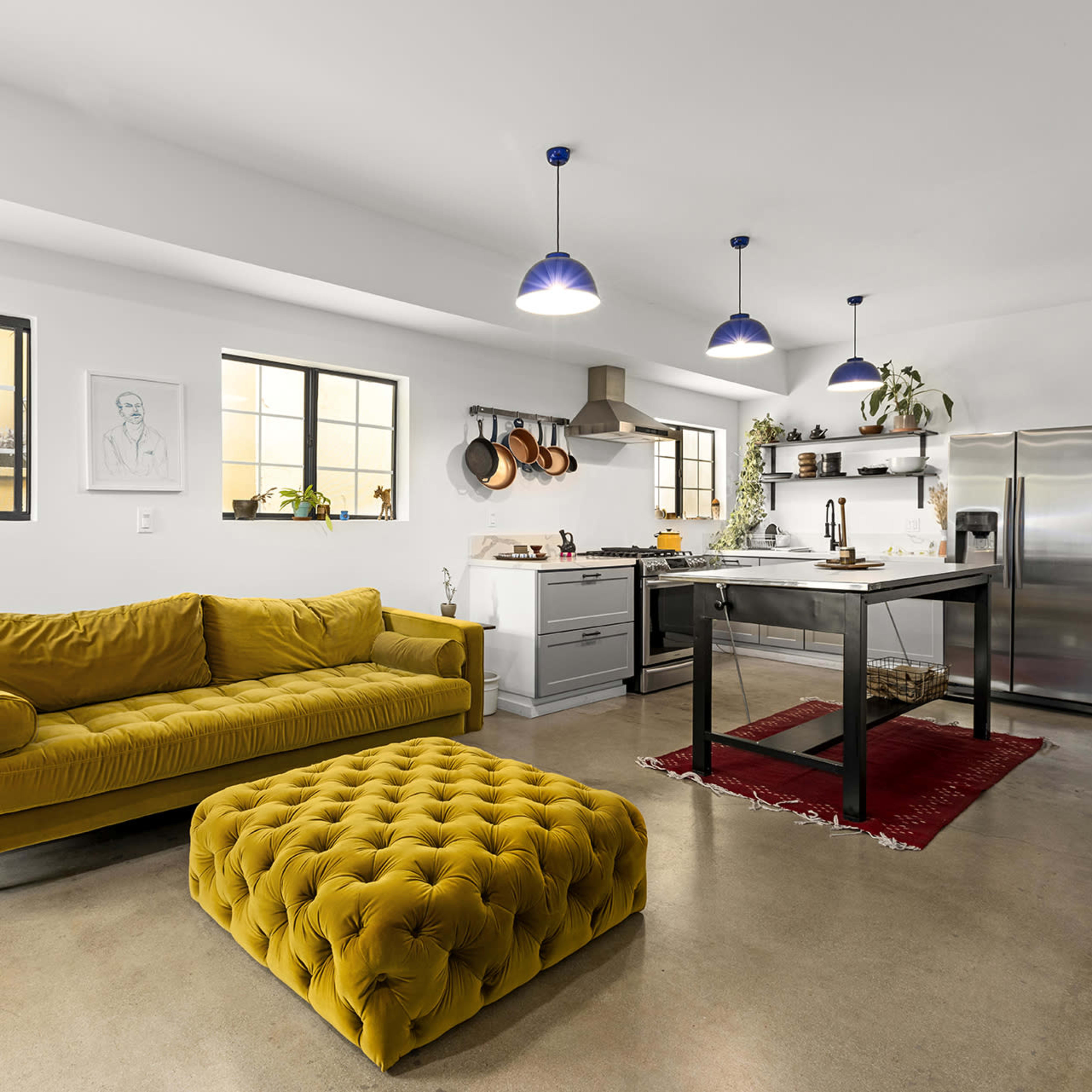 A bright, modern kitchen and living area features a yellow sofa, matching ottoman, and a dining table with industrial-style stools.