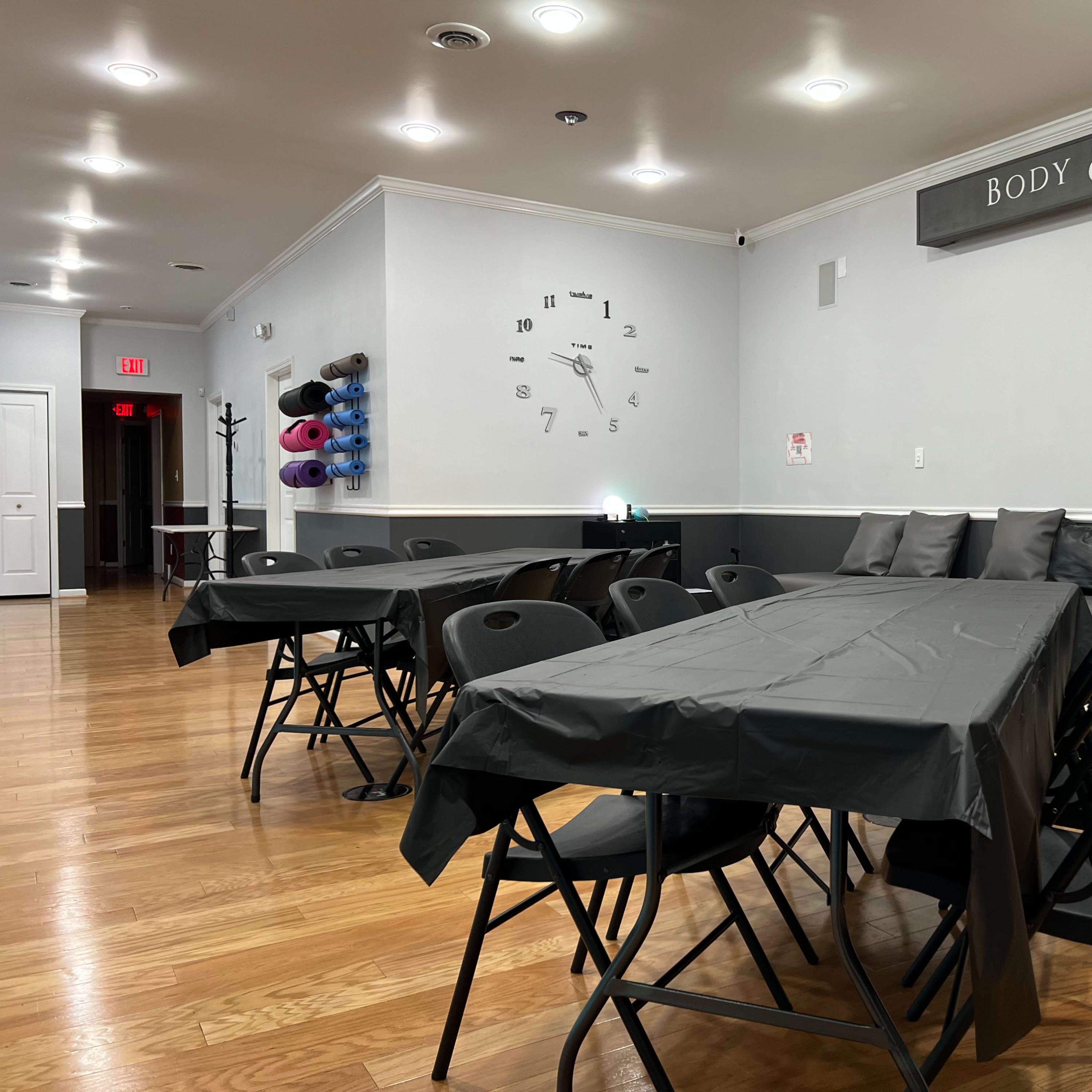 A spacious room with several black folding tables arranged for an event, a wall clock, and a seating area with gray cushions.