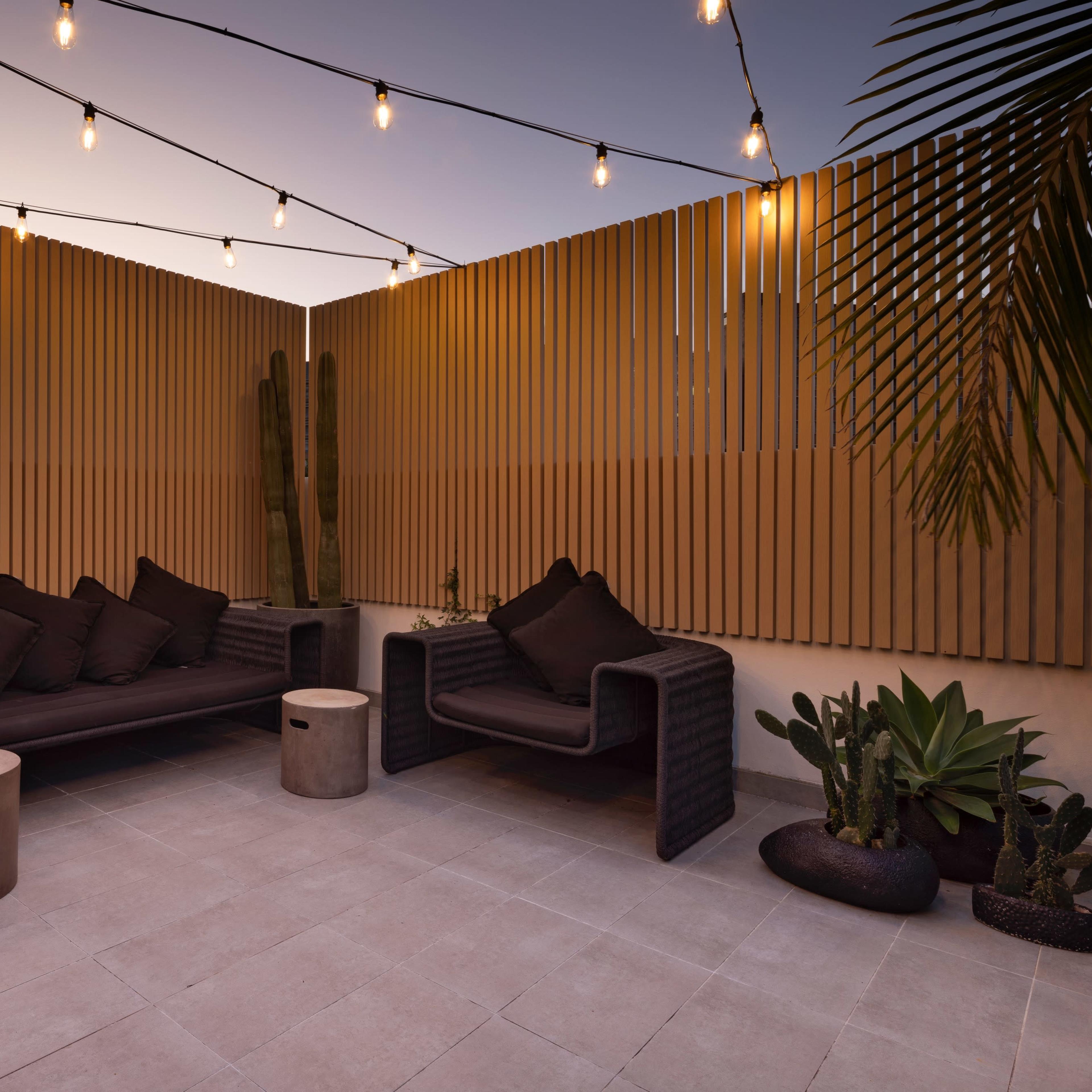 A cozy outdoor seating area with string lights above, surrounded by wooden slats and containing potted plants.