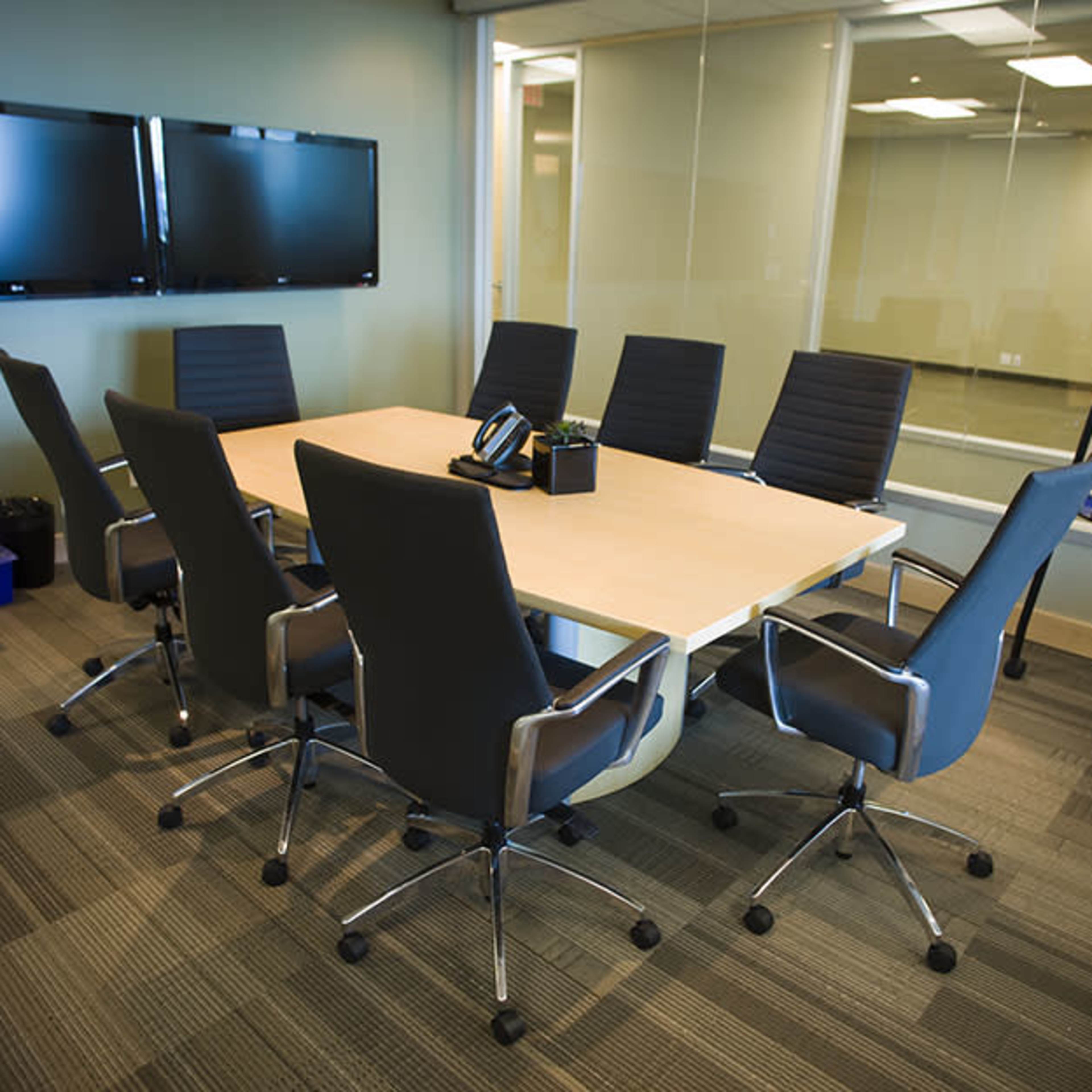 A conference room features a large table surrounded by eight ergonomic chairs, with two wall-mounted screens, a whiteboard, and a phone on the table.