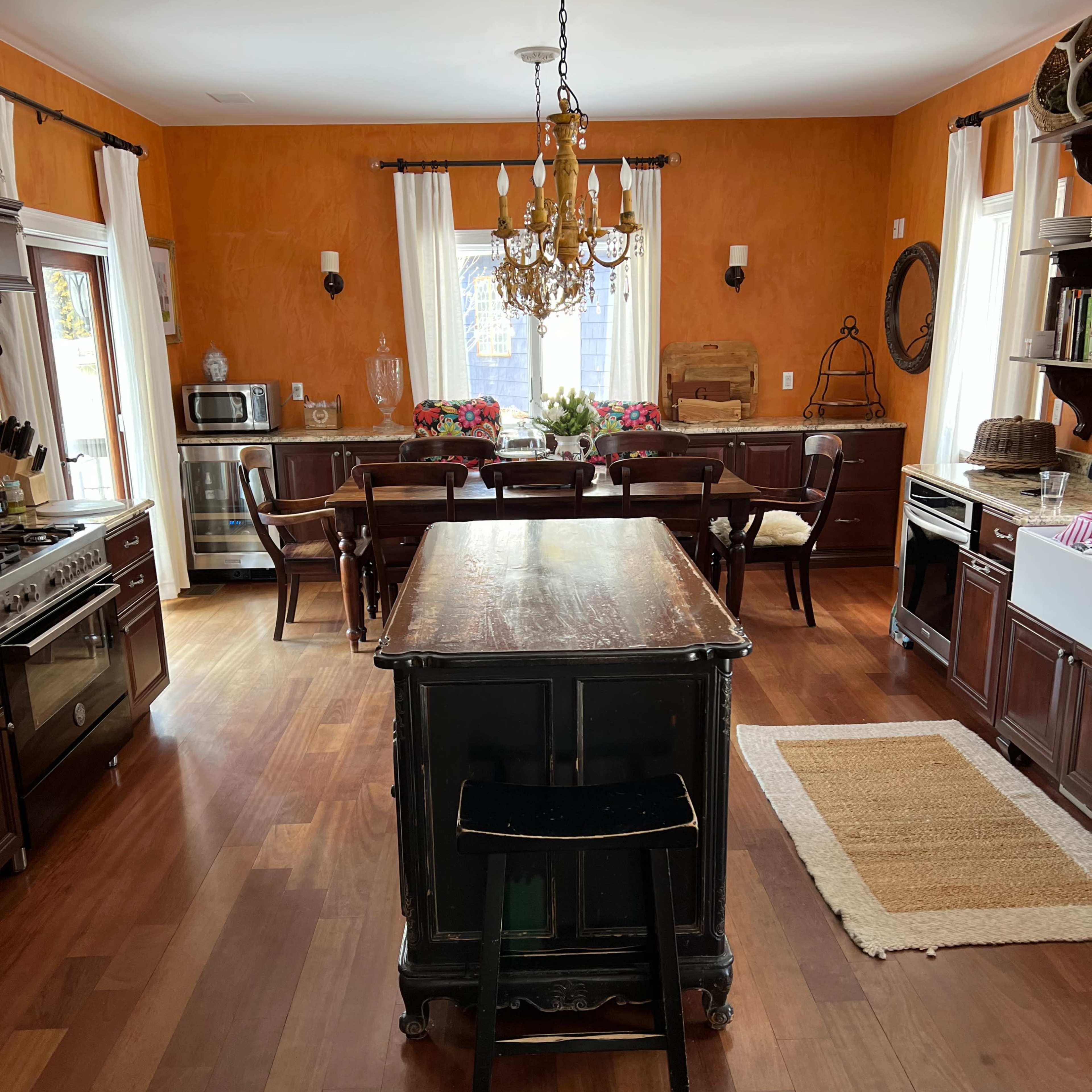 A spacious kitchen with wooden flooring, an island with a black stool, a dining table set with chairs, and orange walls.