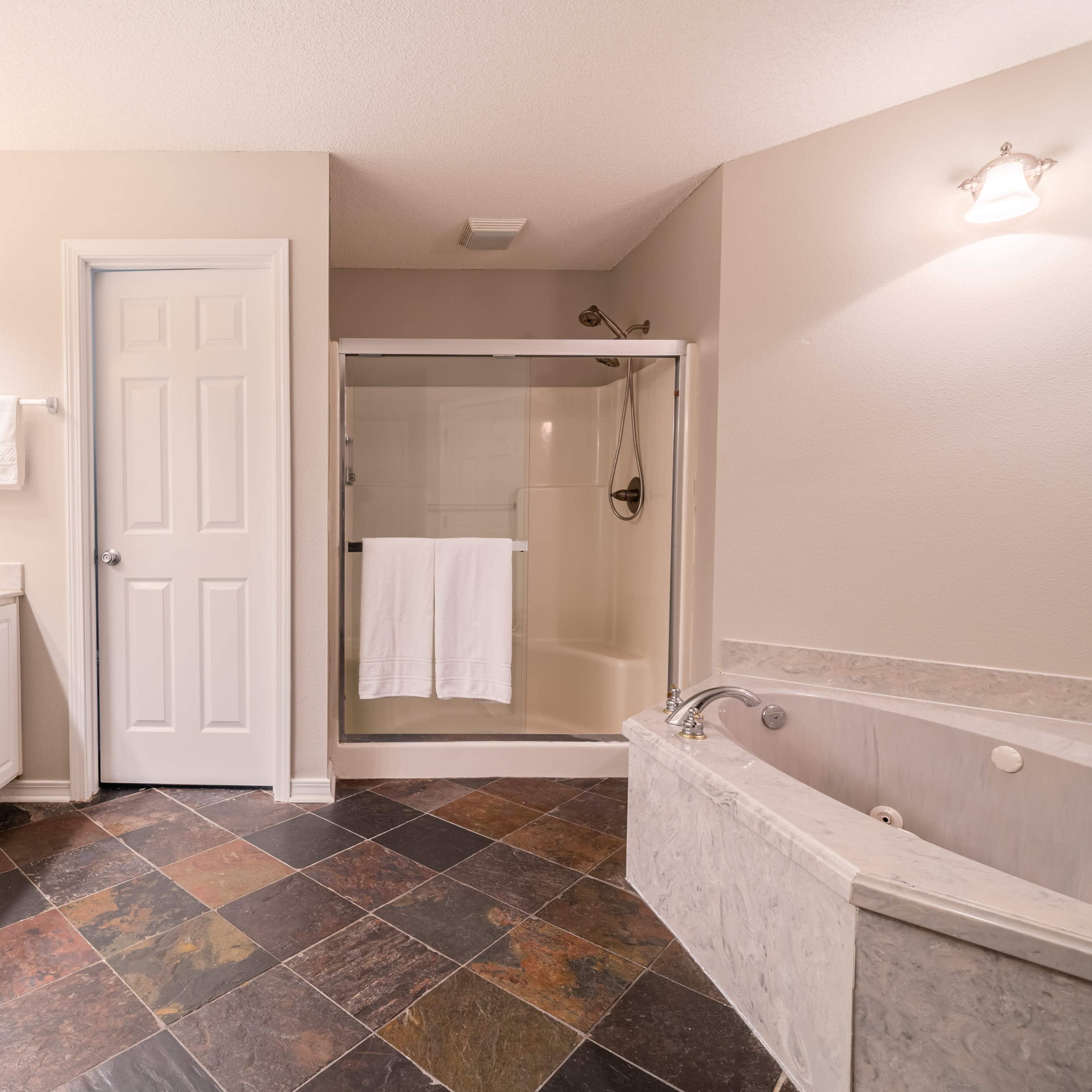 A bathroom features a dual vanity, a glass-enclosed shower, and a jetted tub, with slate flooring and light-colored walls.