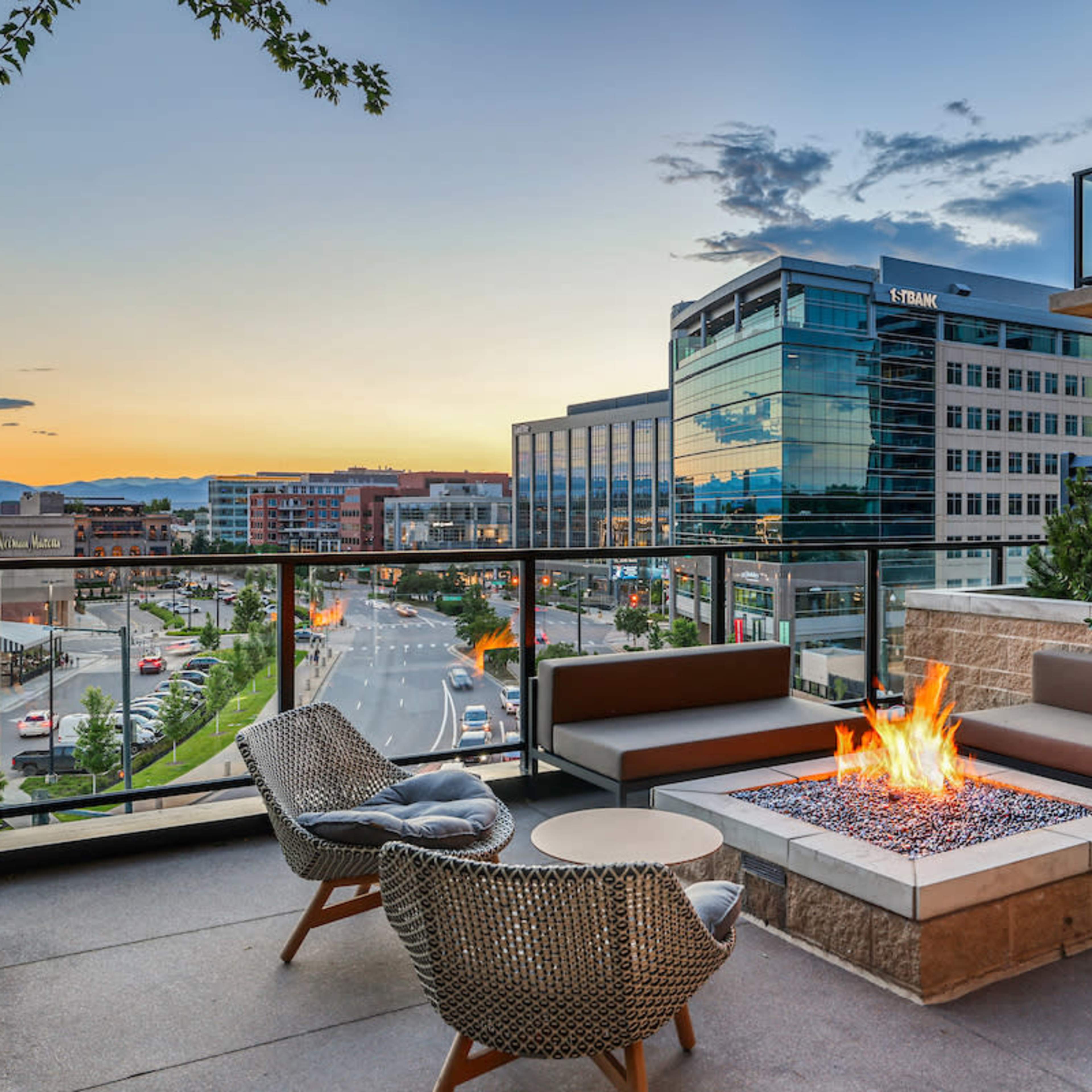 A modern balcony overlooks a city street with a fire pit surrounded by seating, set against a backdrop of buildings and a sunset sky.