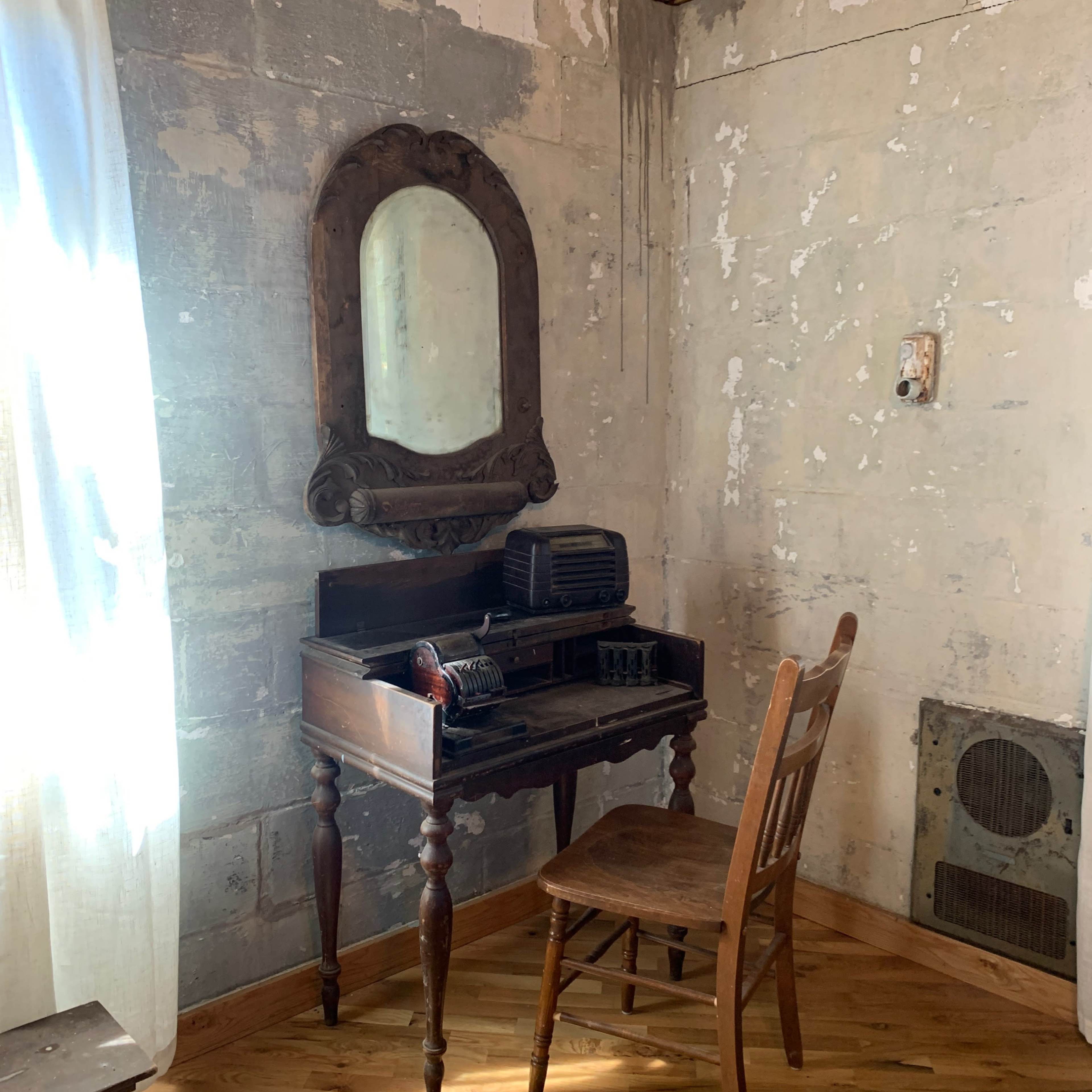 A wooden vanity with a mirror and an old typewriter, accompanied by a wooden chair, set against a wall with peeling paint.