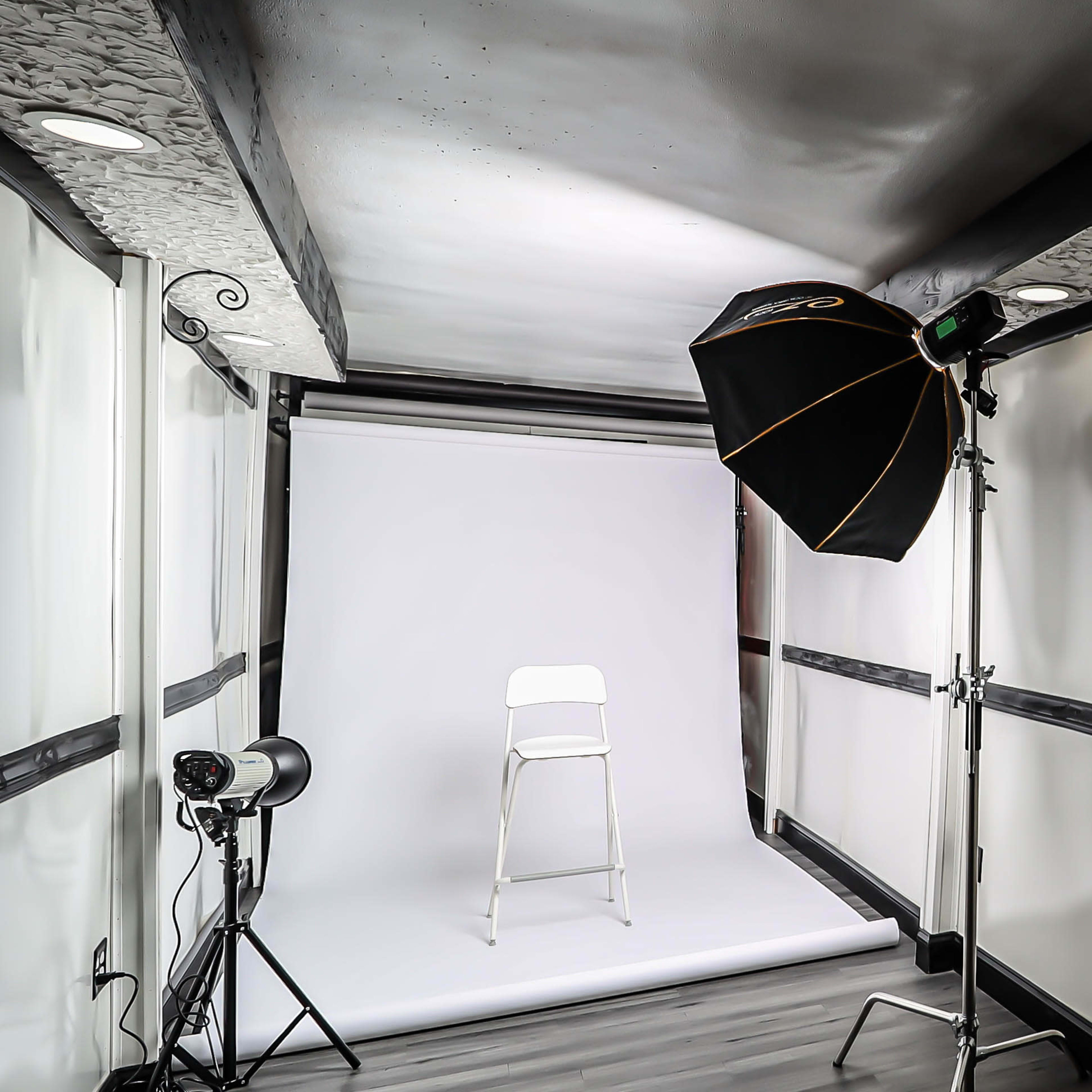 A photography studio features a white backdrop, a single white chair, and lighting equipment set up in a corner.