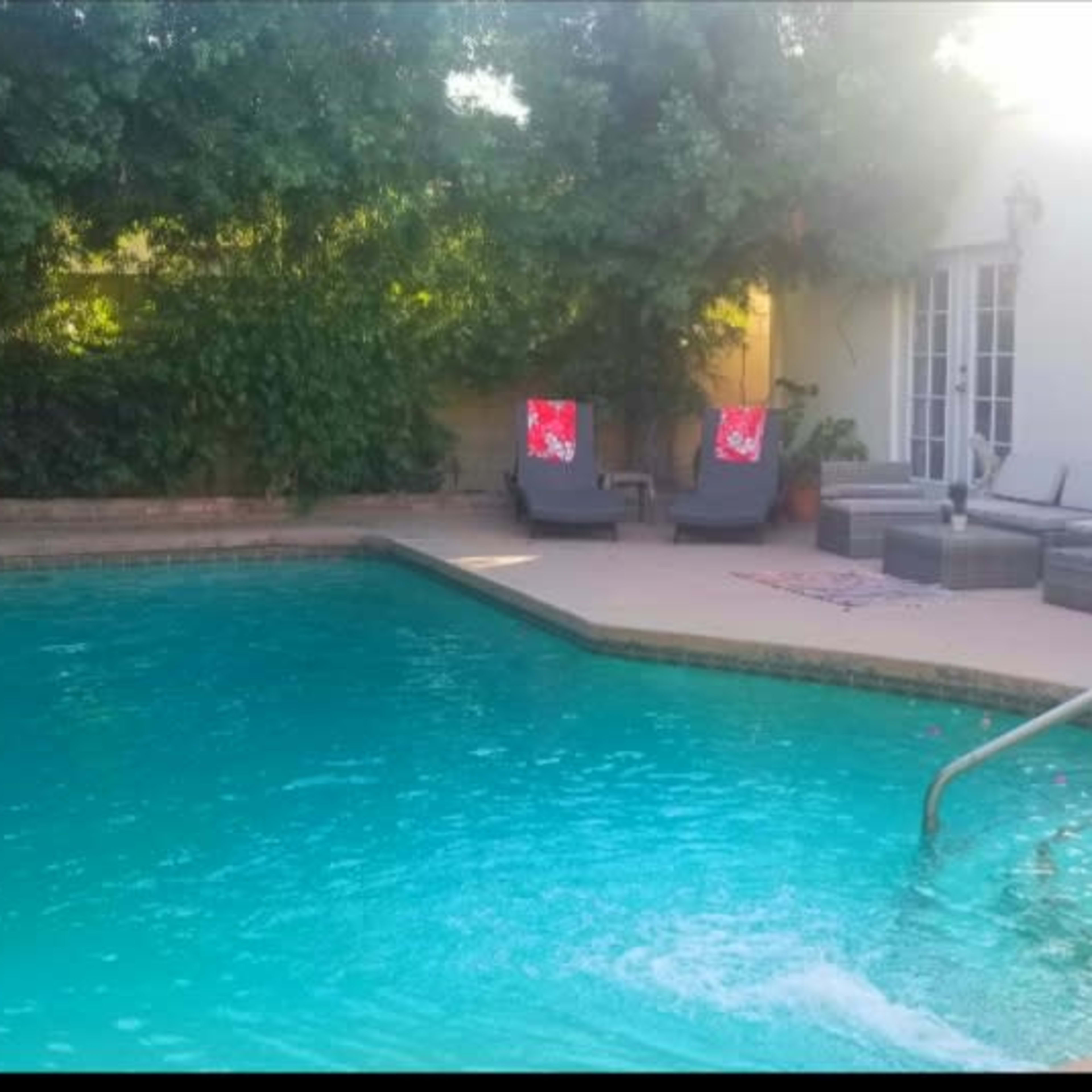 A swimming pool surrounded by green foliage, with lounge chairs and a small table nearby.