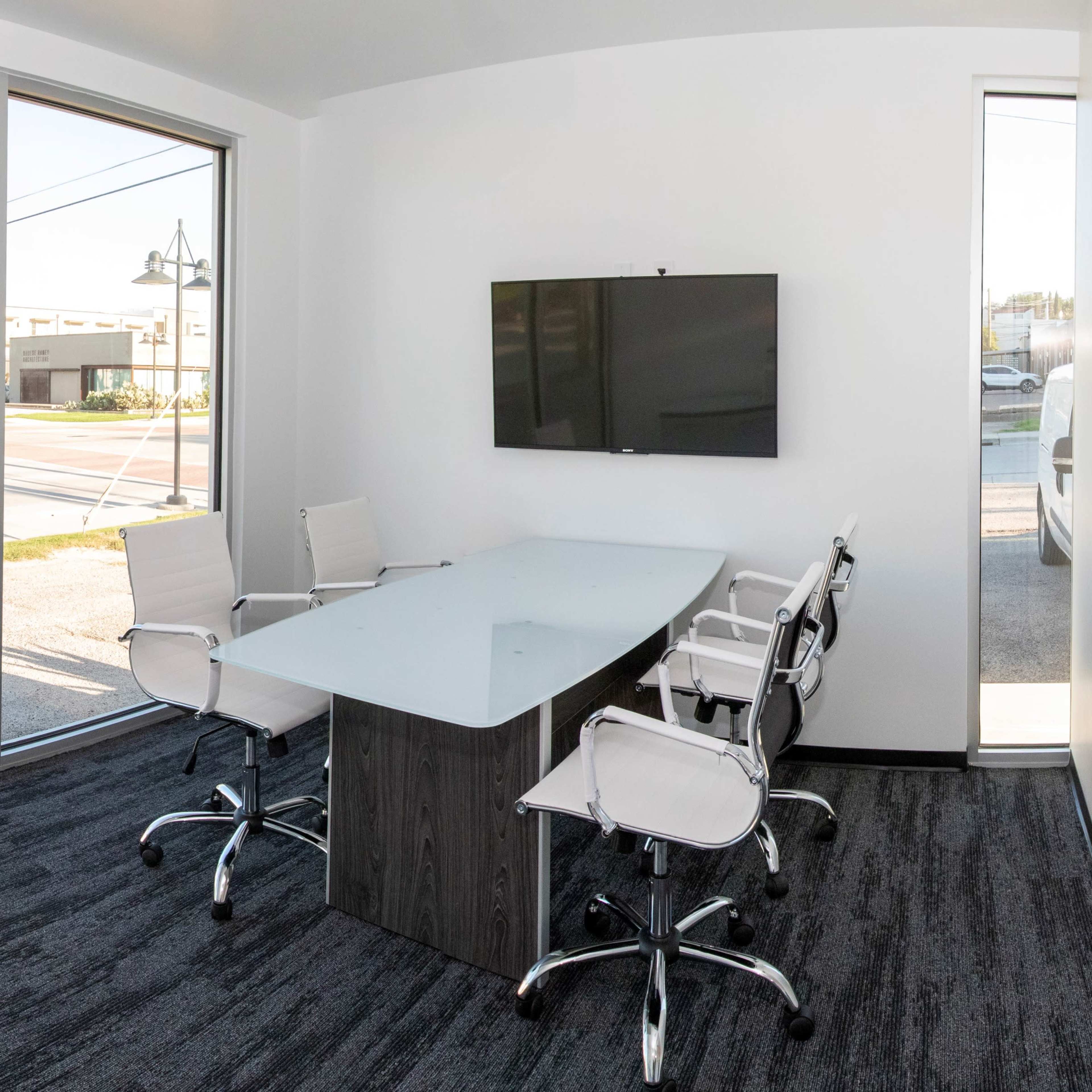 A modern conference room with a glass wall, a rectangular table, and white chairs, featuring a television on the wall.