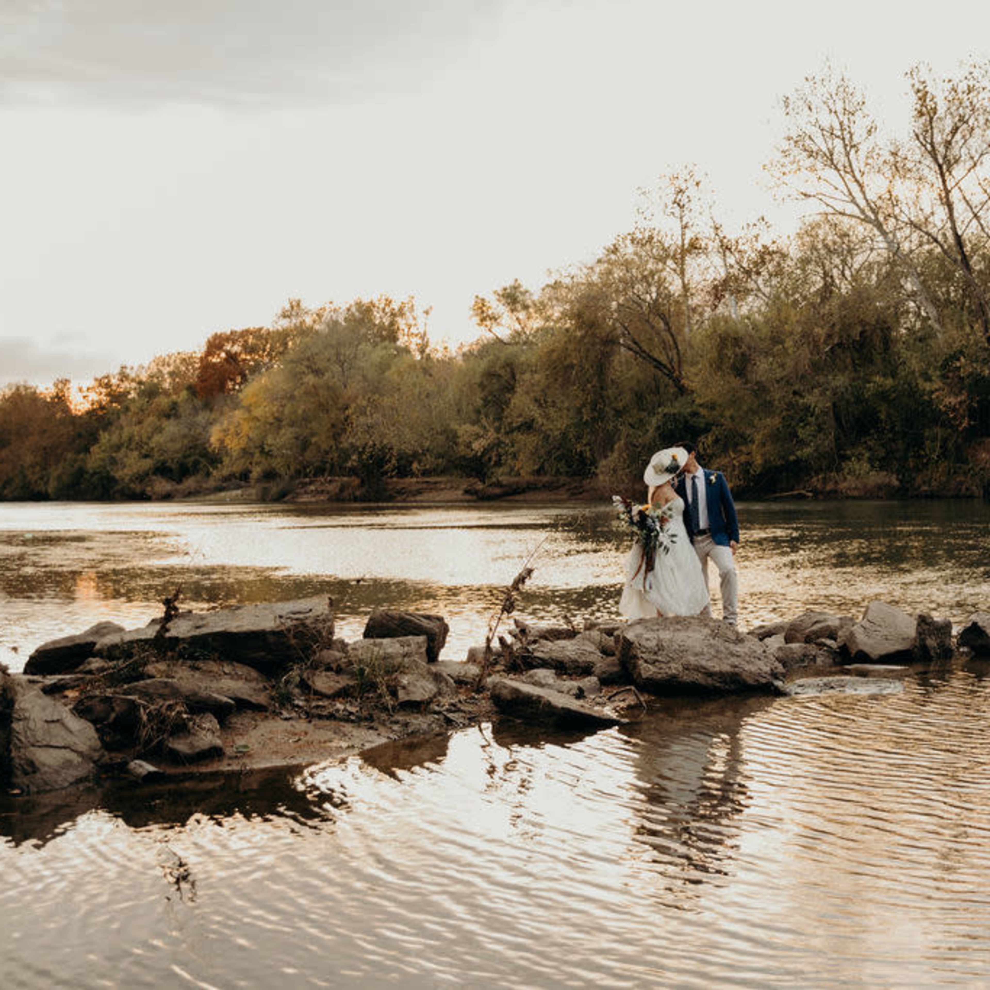 A couple in wedding attire stands on large rocks in a river, surrounded by trees as the sun sets in the background.
