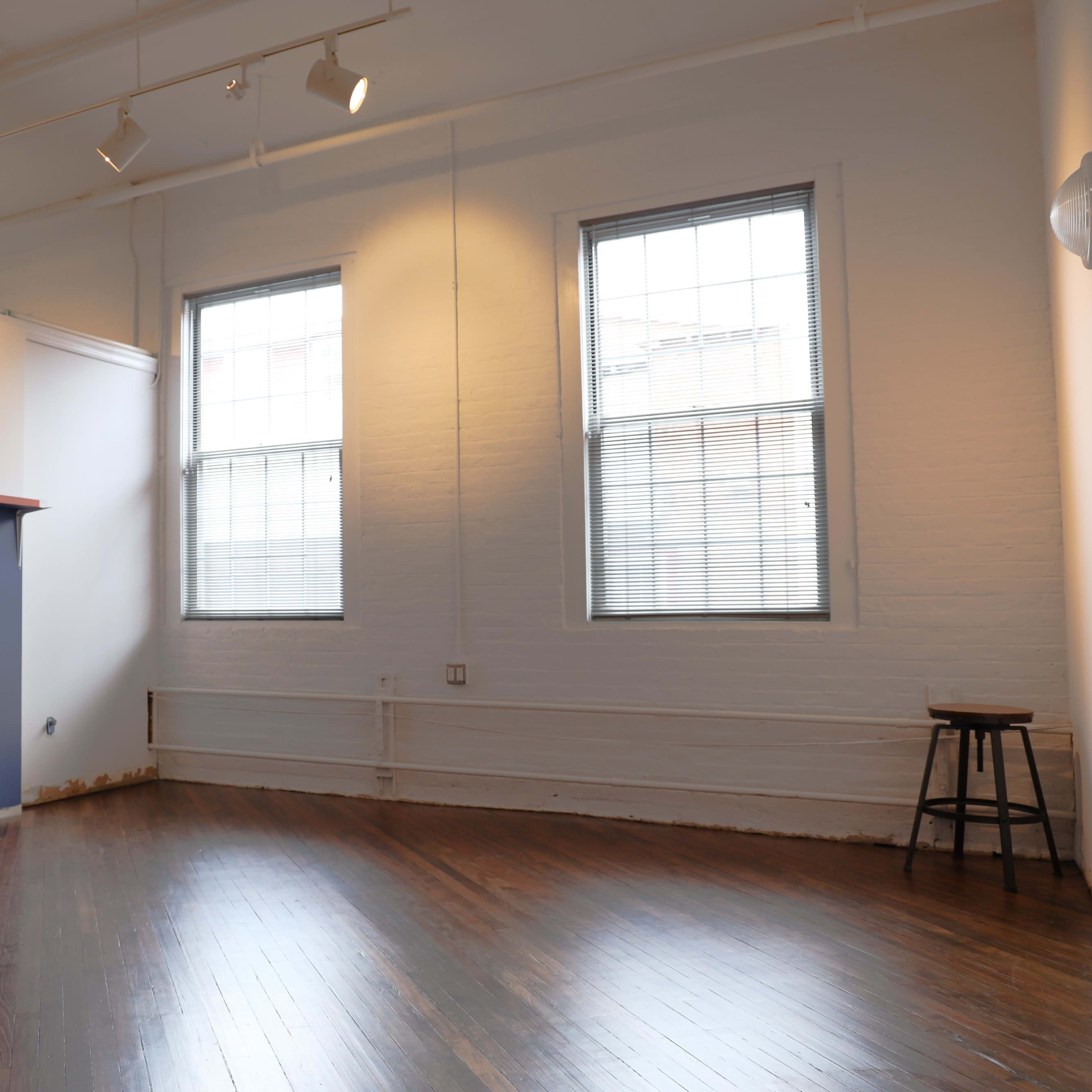 An empty room with hardwood floors, two windows, a blue wall section, and a wooden stool in the corner.