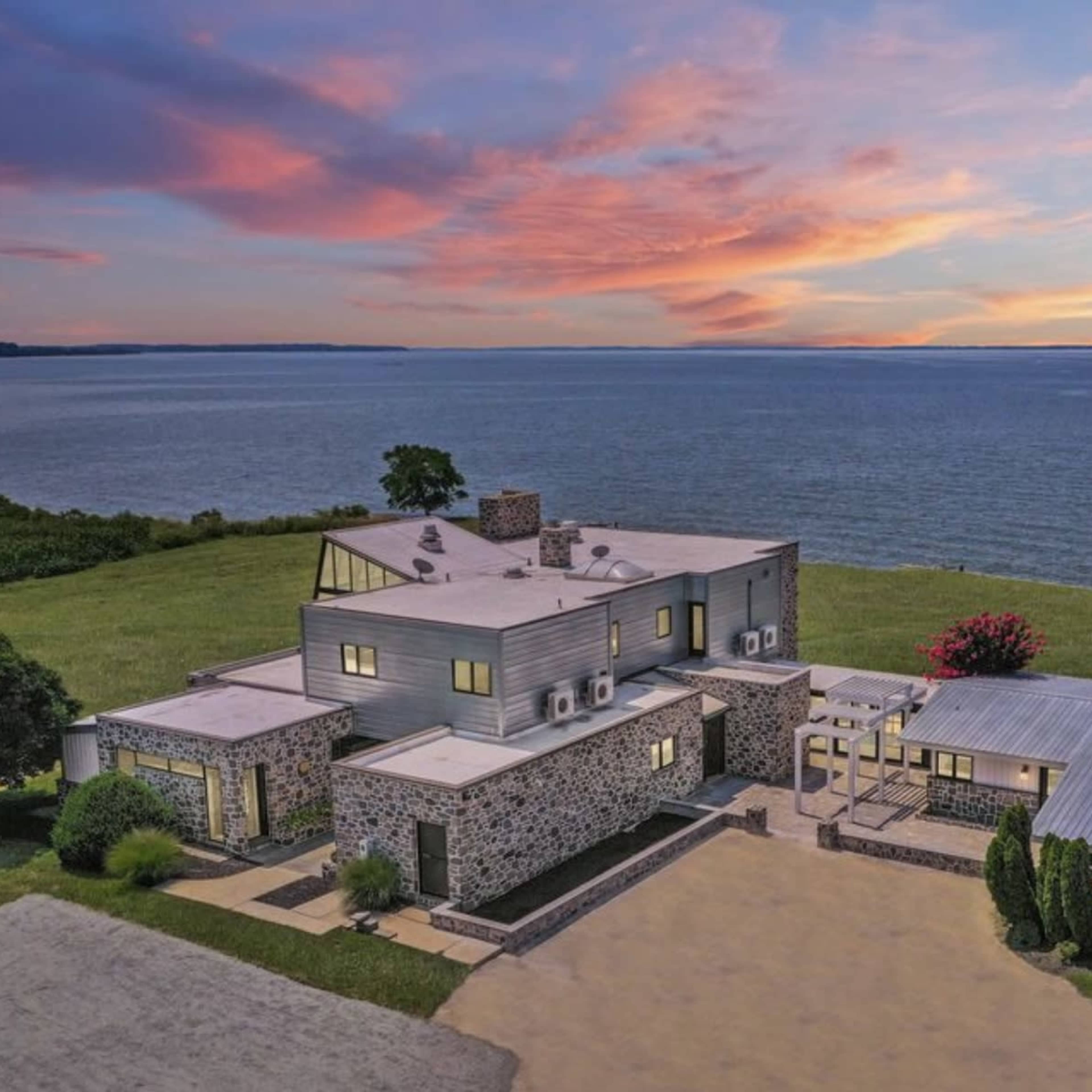 A modern stone and metal house sits on a grassy shoreline overlooking a body of water at sunset.