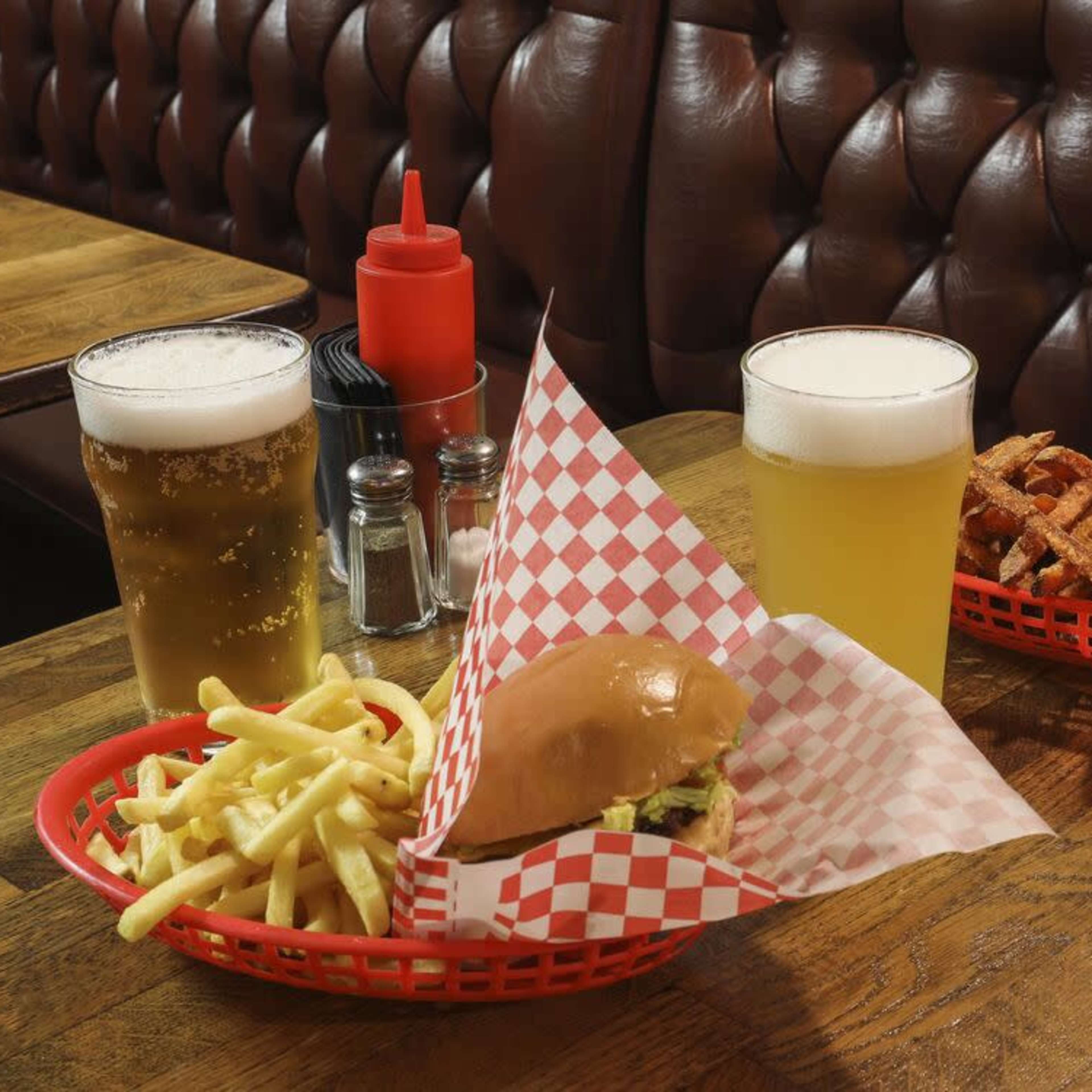Two baskets of burgers and fries, along with three glasses of beer, are placed on a wooden table in a cushioned booth setting.