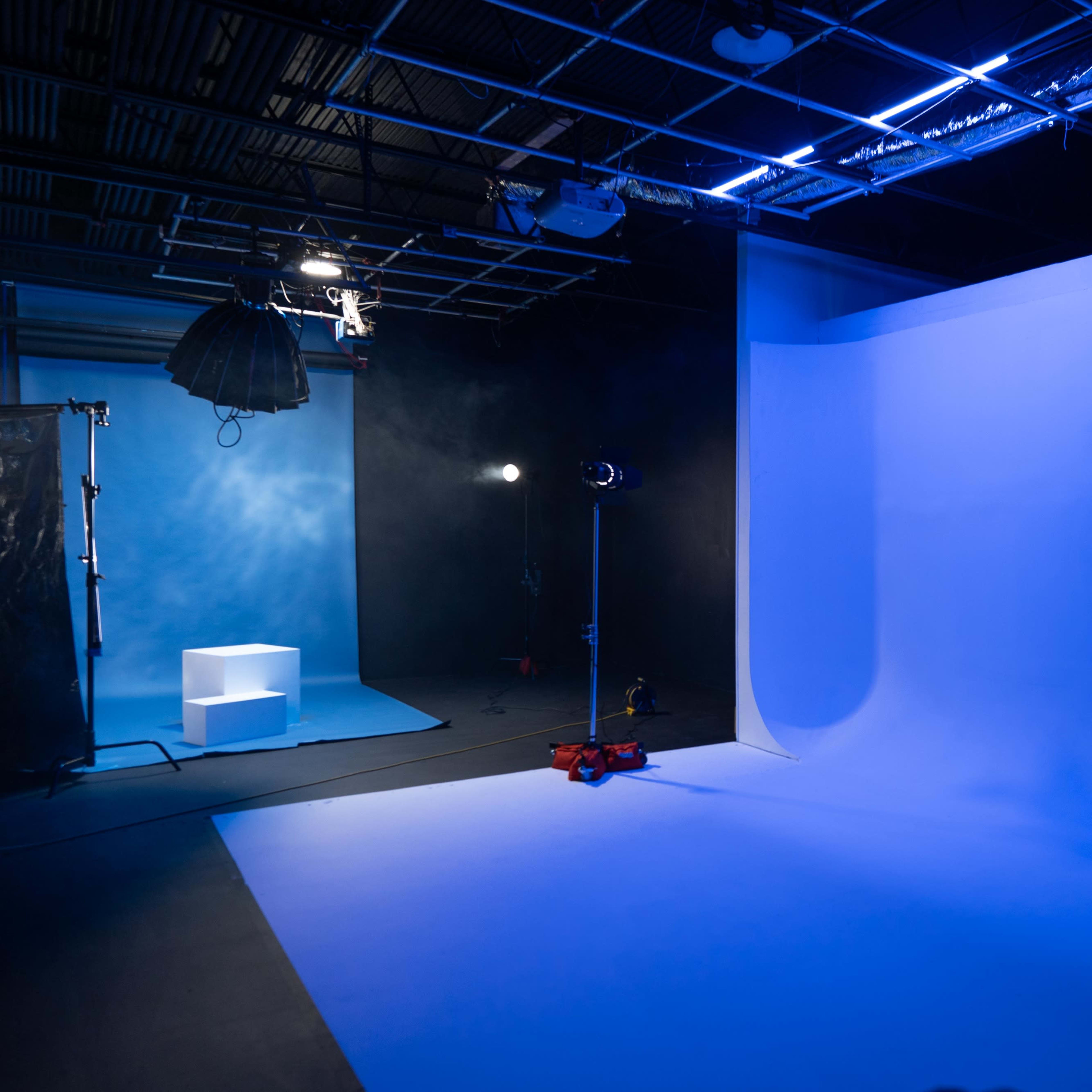 A photography studio with a blue backdrop, a light setup on the left, and a small white platform in the background.