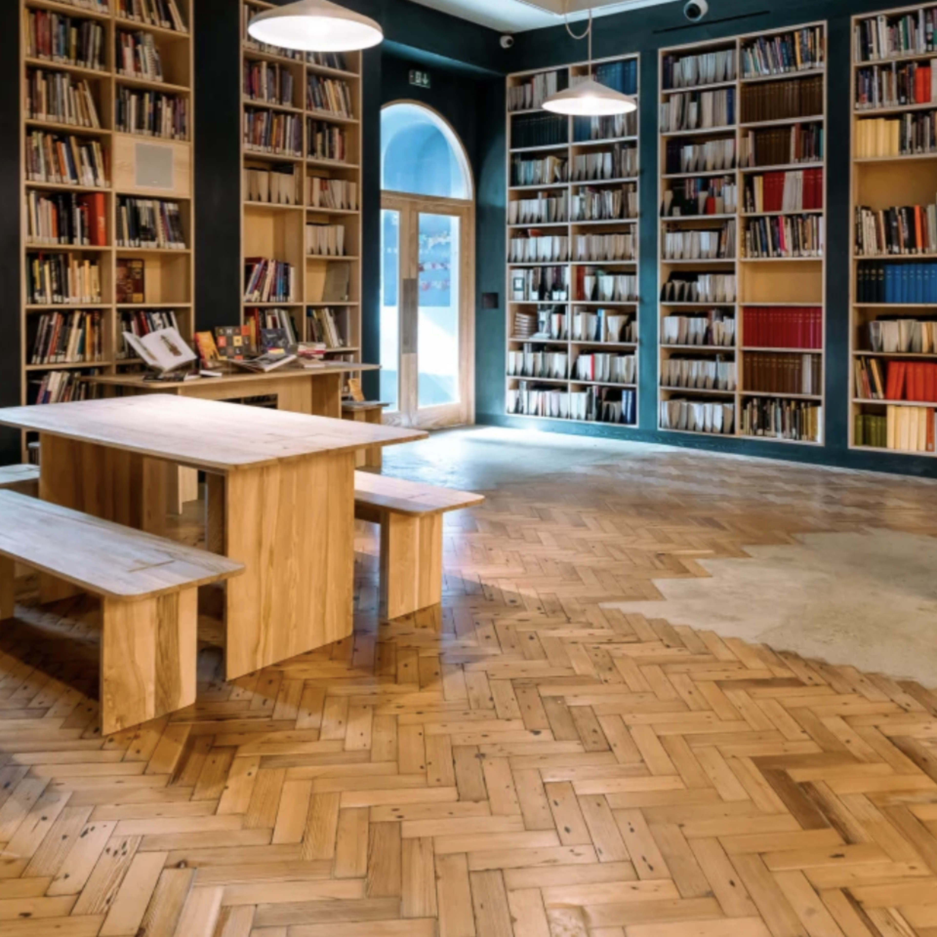 A modern library interior featuring tall shelves filled with books, a wooden table with benches, and a herringbone pattern wooden floor.