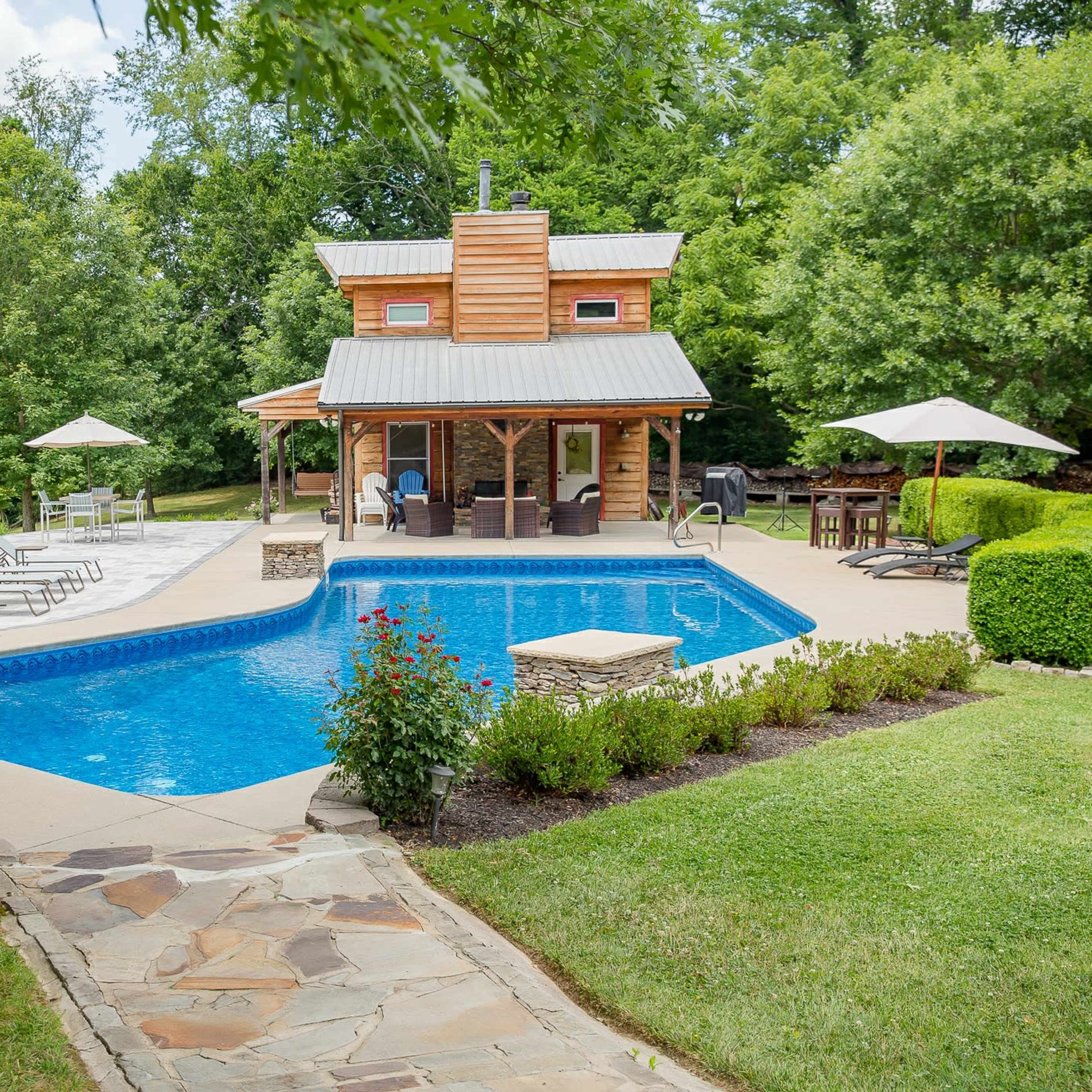 A wooden cabin beside a rectangular swimming pool with lounge chairs and tables under umbrellas, set among green trees and a grassy area.