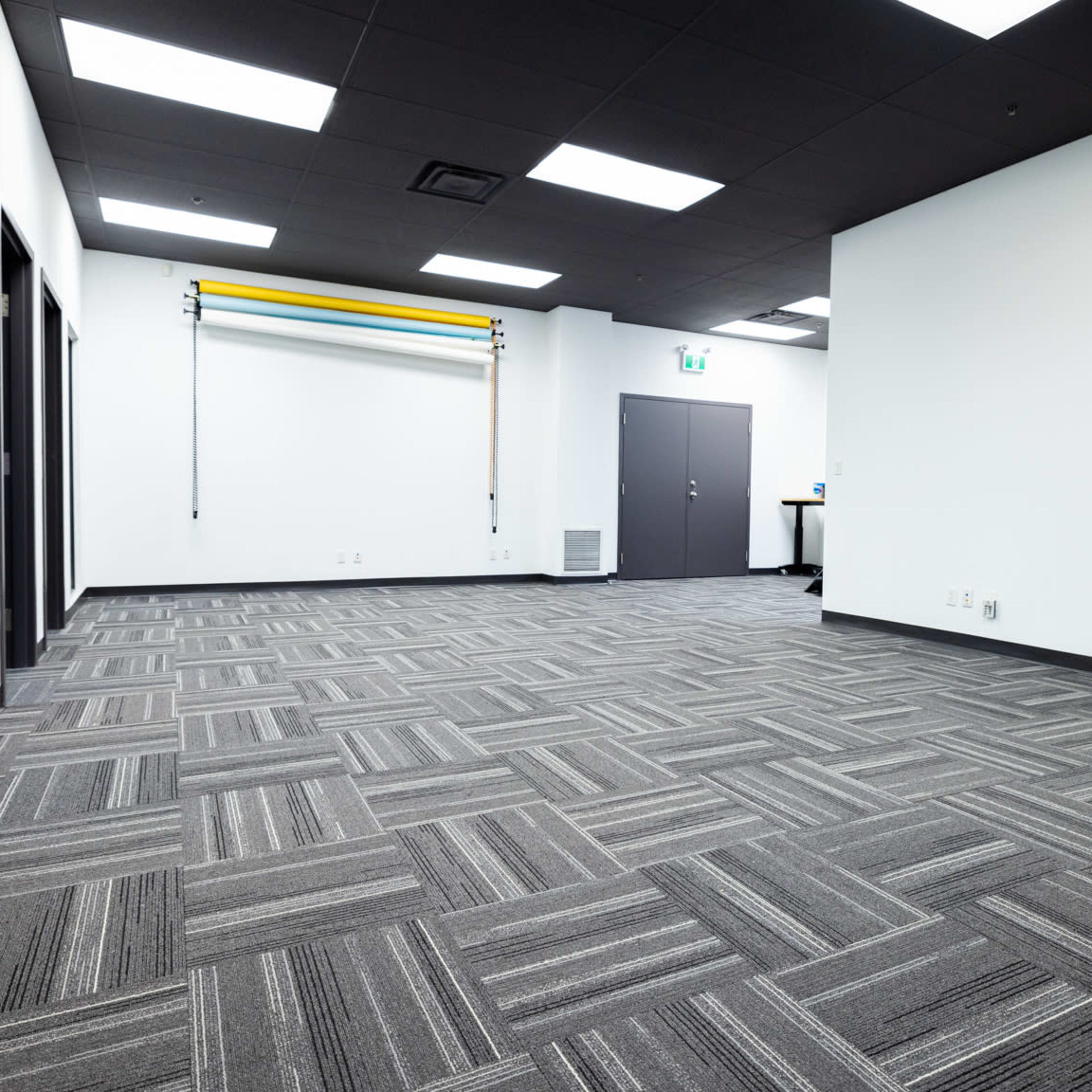 A large, empty room with gray carpeting, white walls, and overhead fluorescent lighting, featuring mirrors and a closed door on one side.