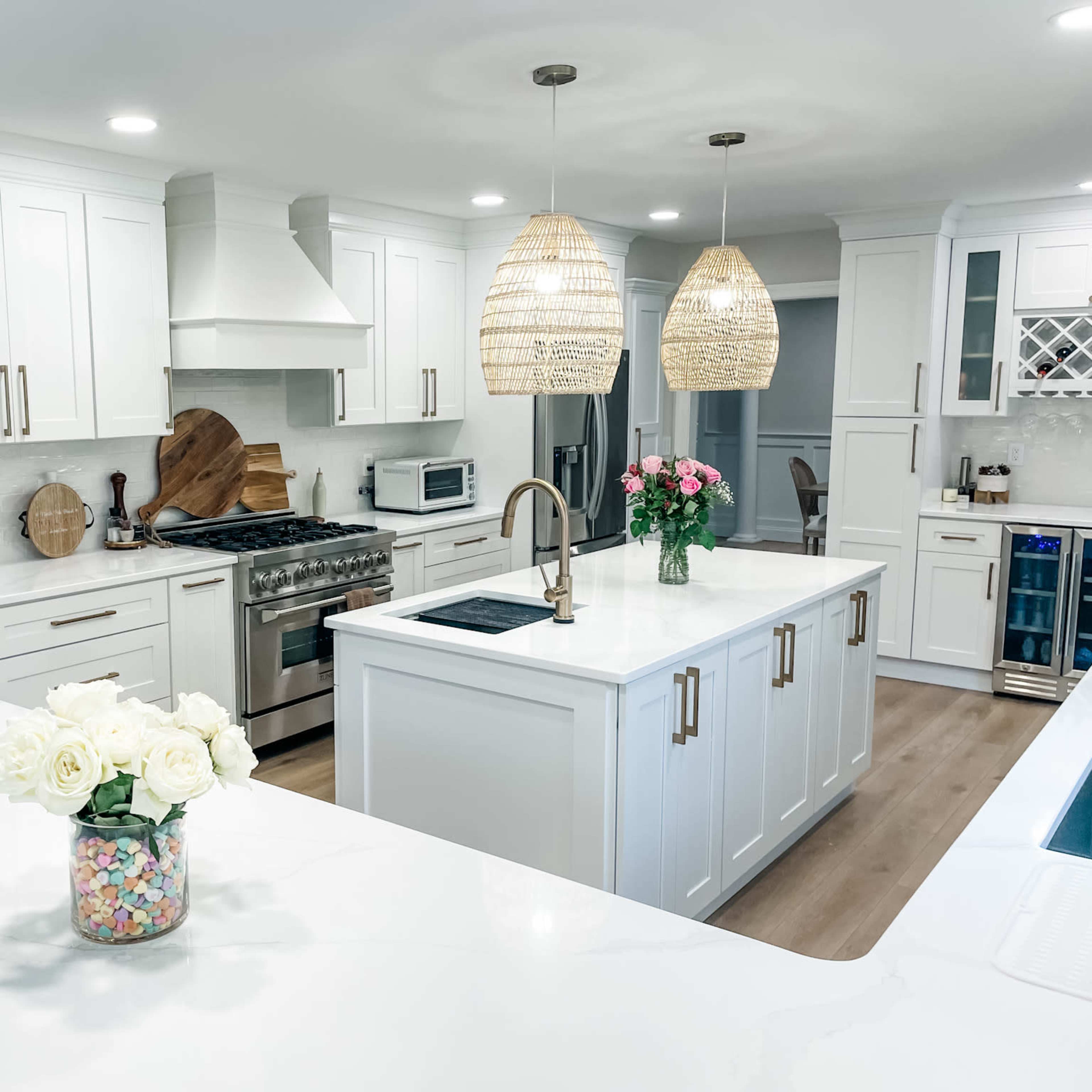 A modern kitchen features white cabinetry, a large island with a sink, and pendant lighting above, complemented by freshly cut flowers in a jar on the counter.