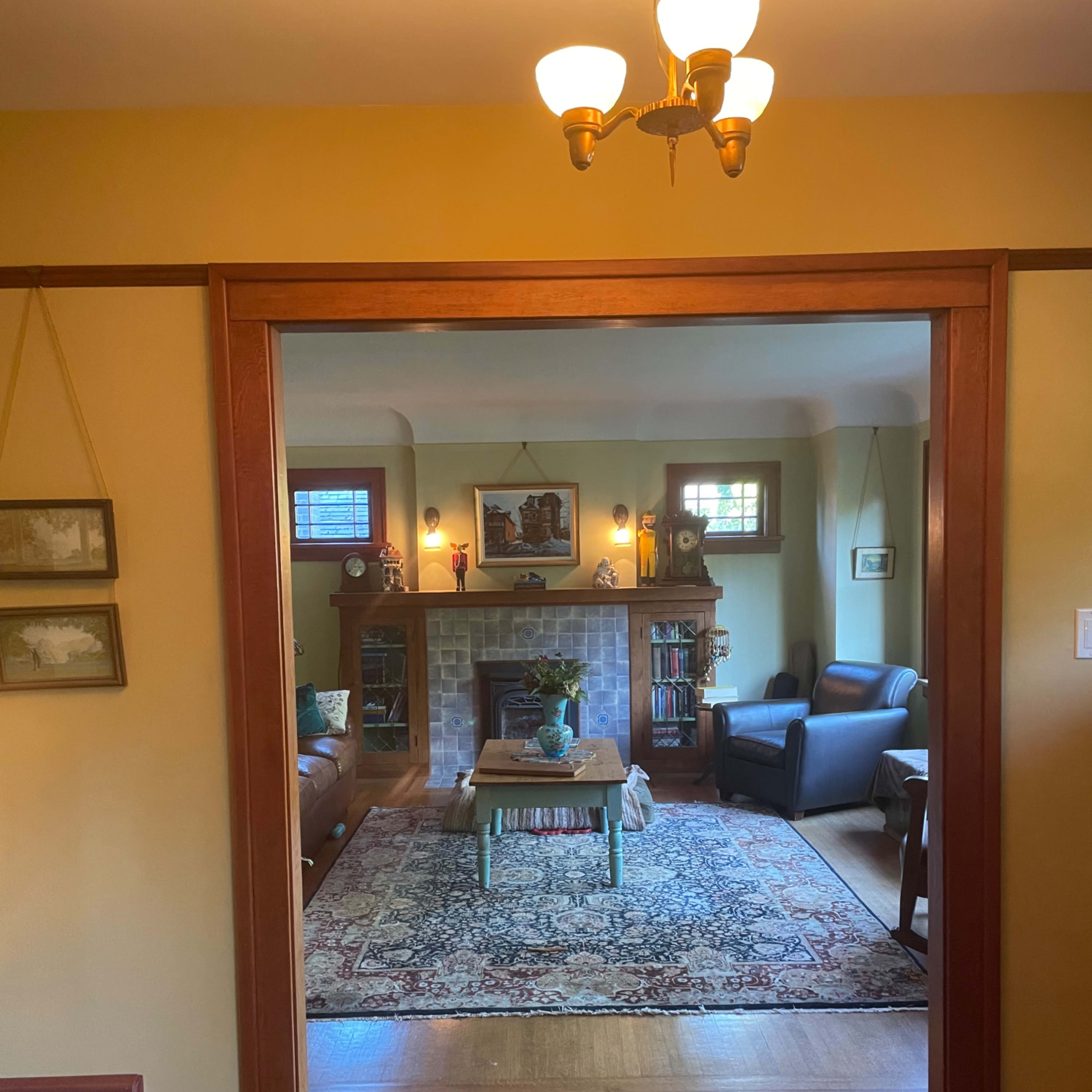 A cozy living room is framed by an open doorway, featuring a fireplace, a coffee table, and upholstered seating around a decorative rug.