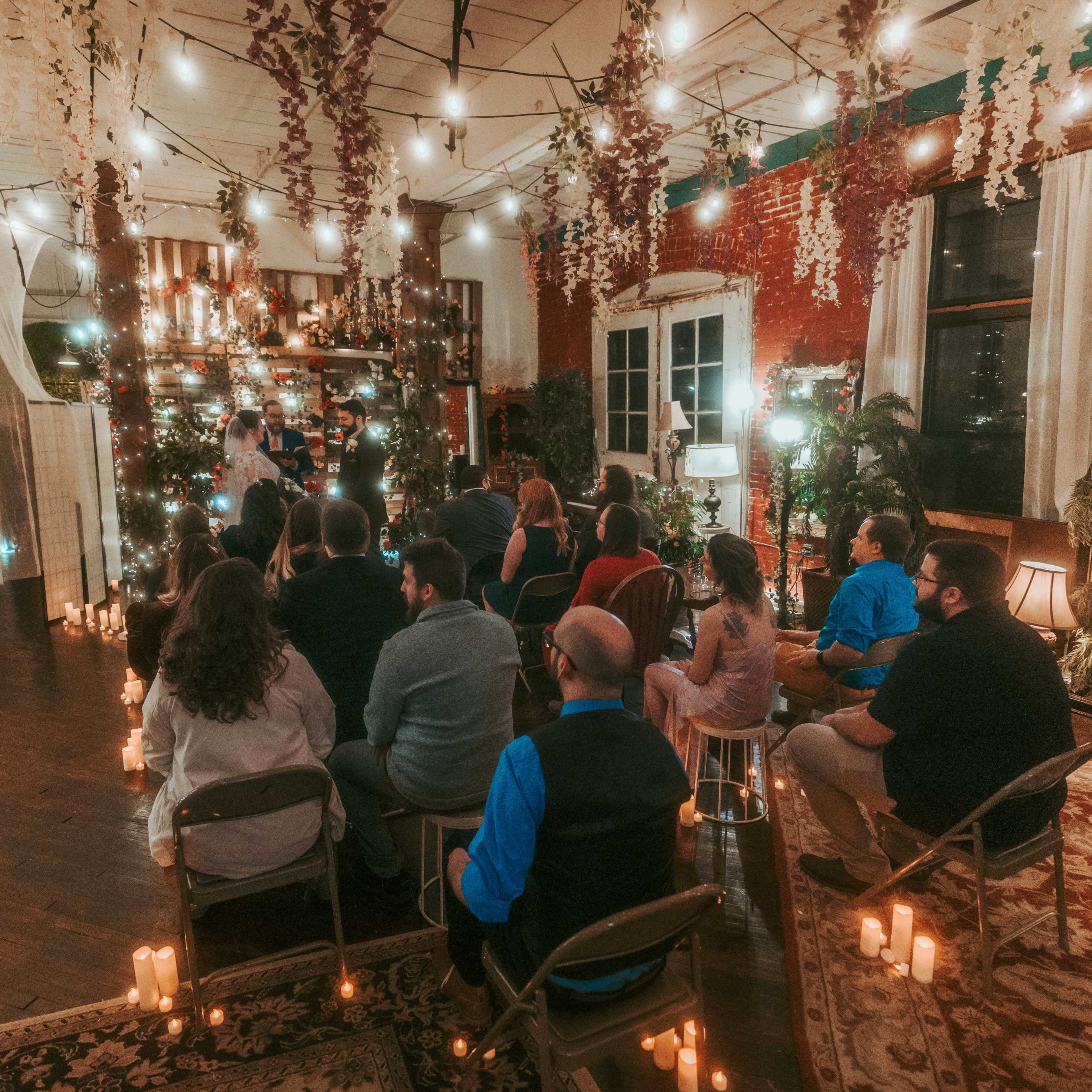 A group of people sits in chairs facing a decorated altar illuminated by string lights and candles in a vintage indoor space.