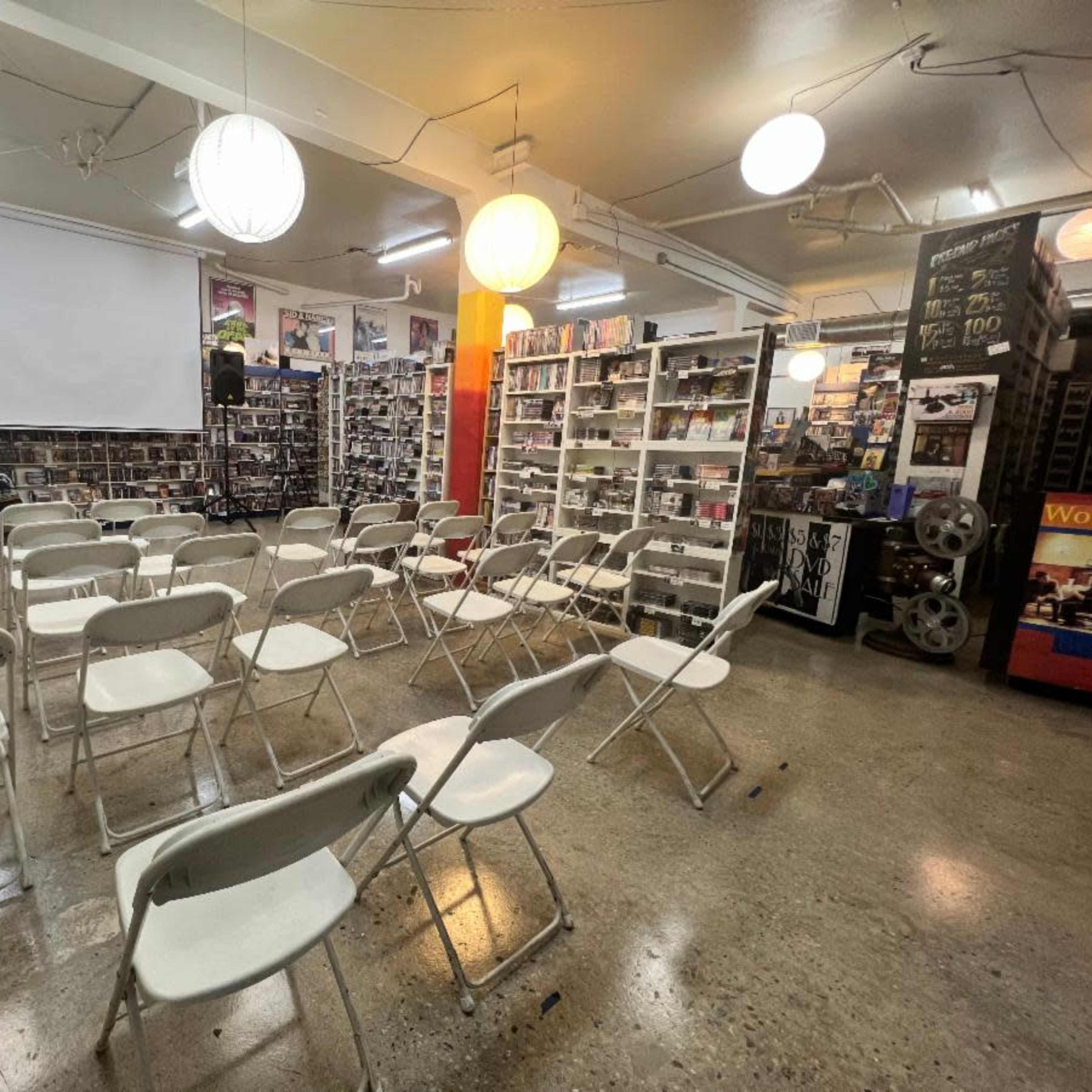 A room arranged with rows of white folding chairs facing a blank projector screen, surrounded by shelves filled with DVDs and other media.