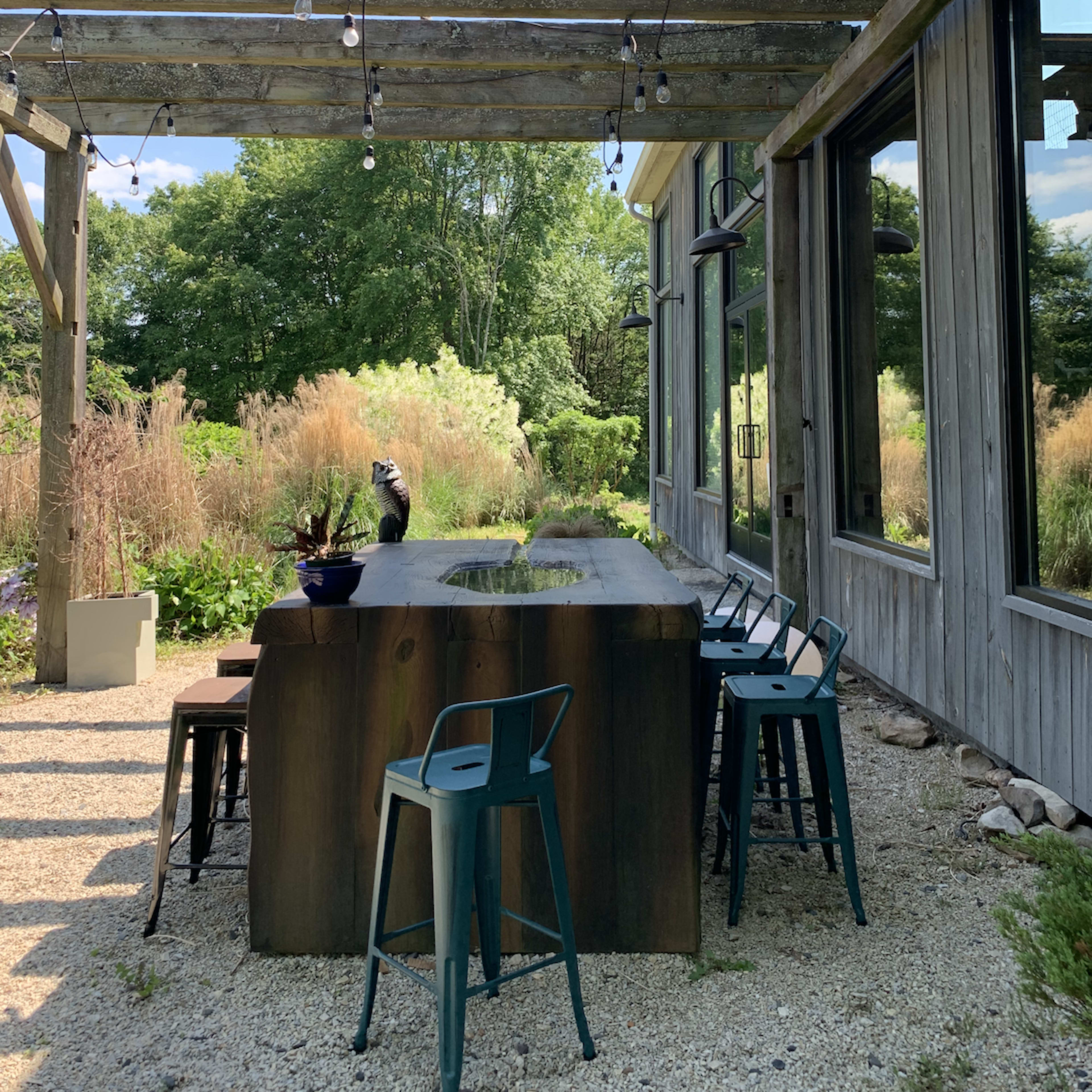 A wooden bar with metal stools is situated under a wooden pergola adorned with string lights, surrounded by a garden.