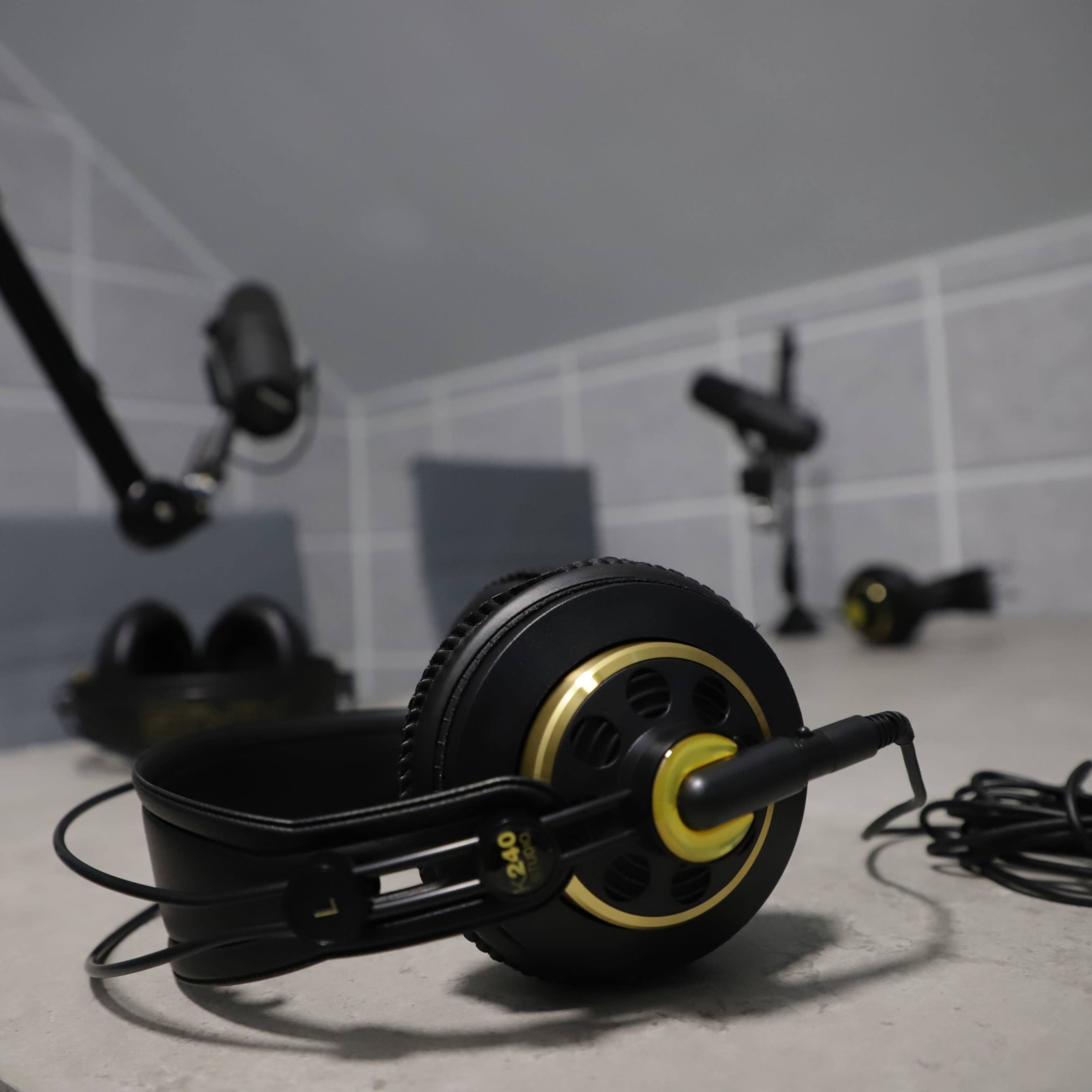 A pair of black headphones with gold accents sits on a concrete surface, surrounded by various microphones and audio equipment in a minimalist workspace.