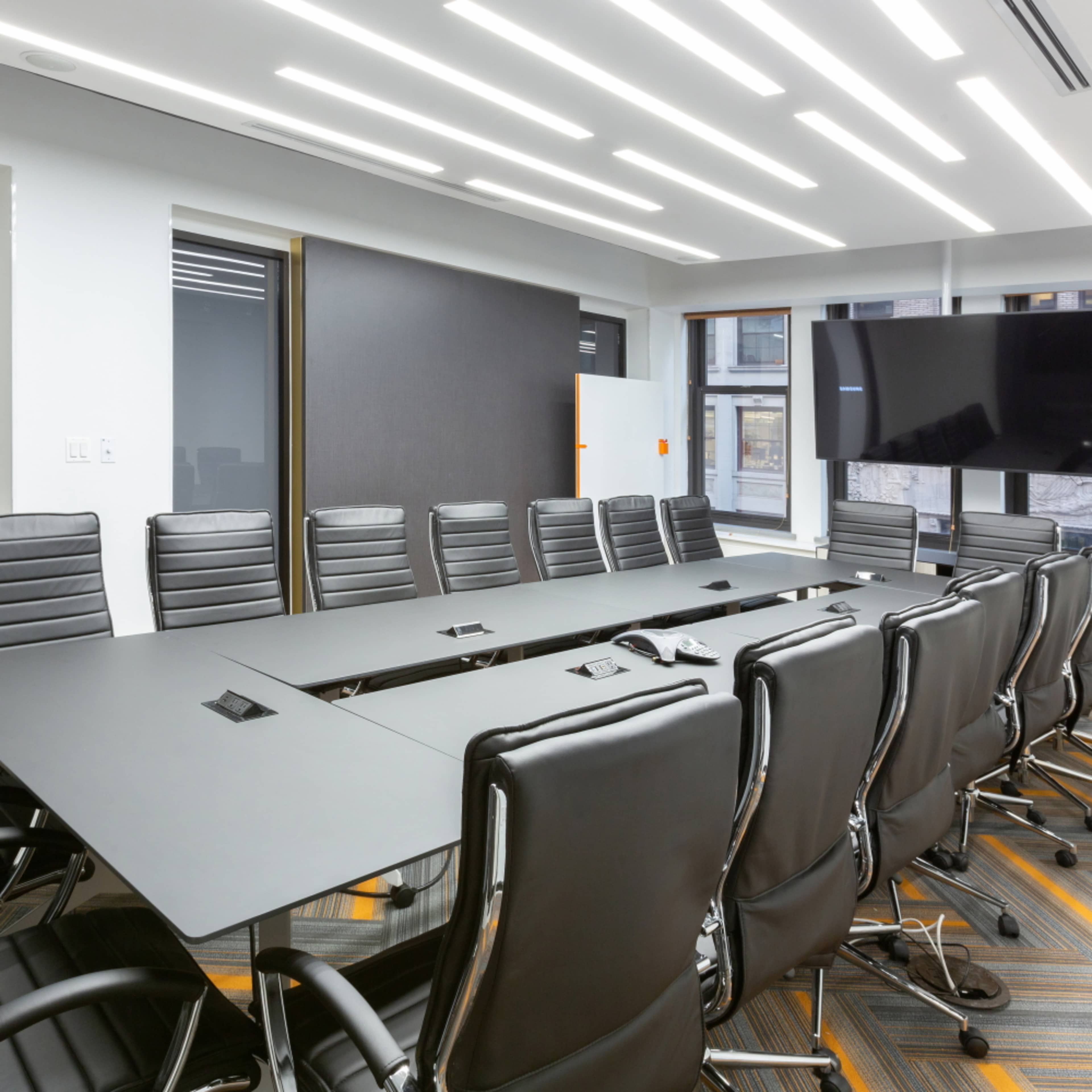 A modern conference room features a long black table surrounded by multiple black ergonomic chairs, with a large television mounted on the wall and illuminated by overhead LED lights.