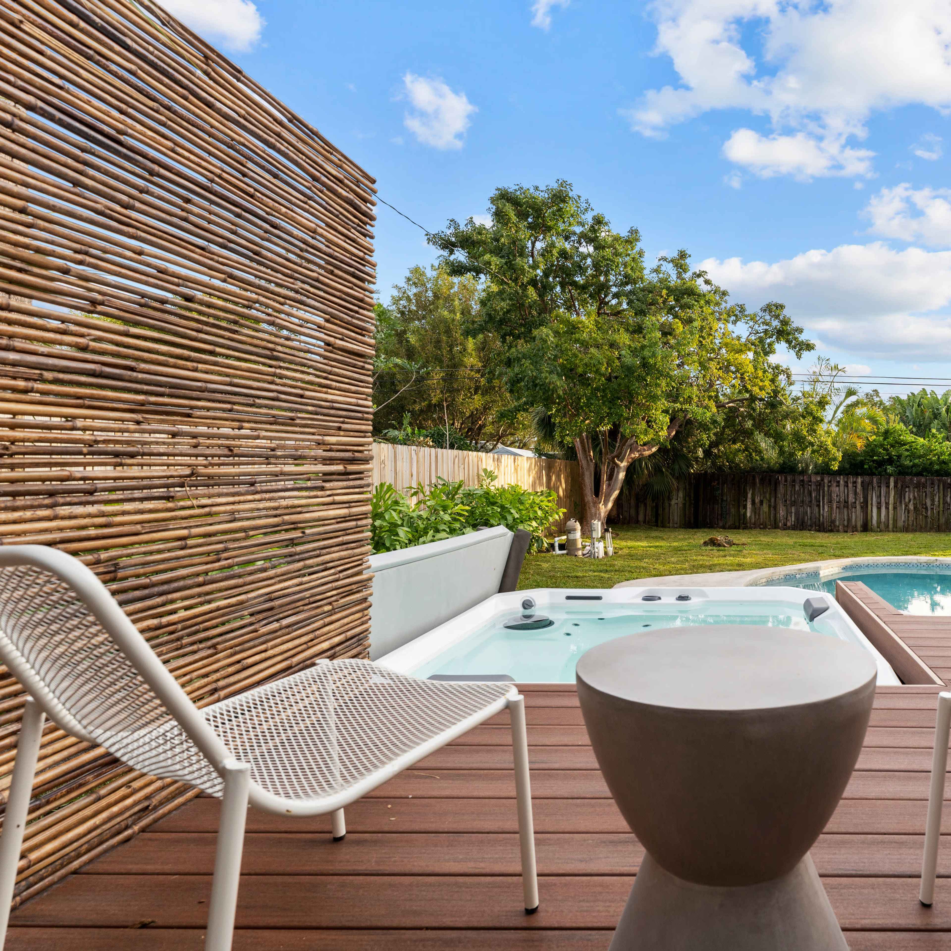 A patio features two white chairs and a round table overlooking a swimming pool surrounded by greenery.