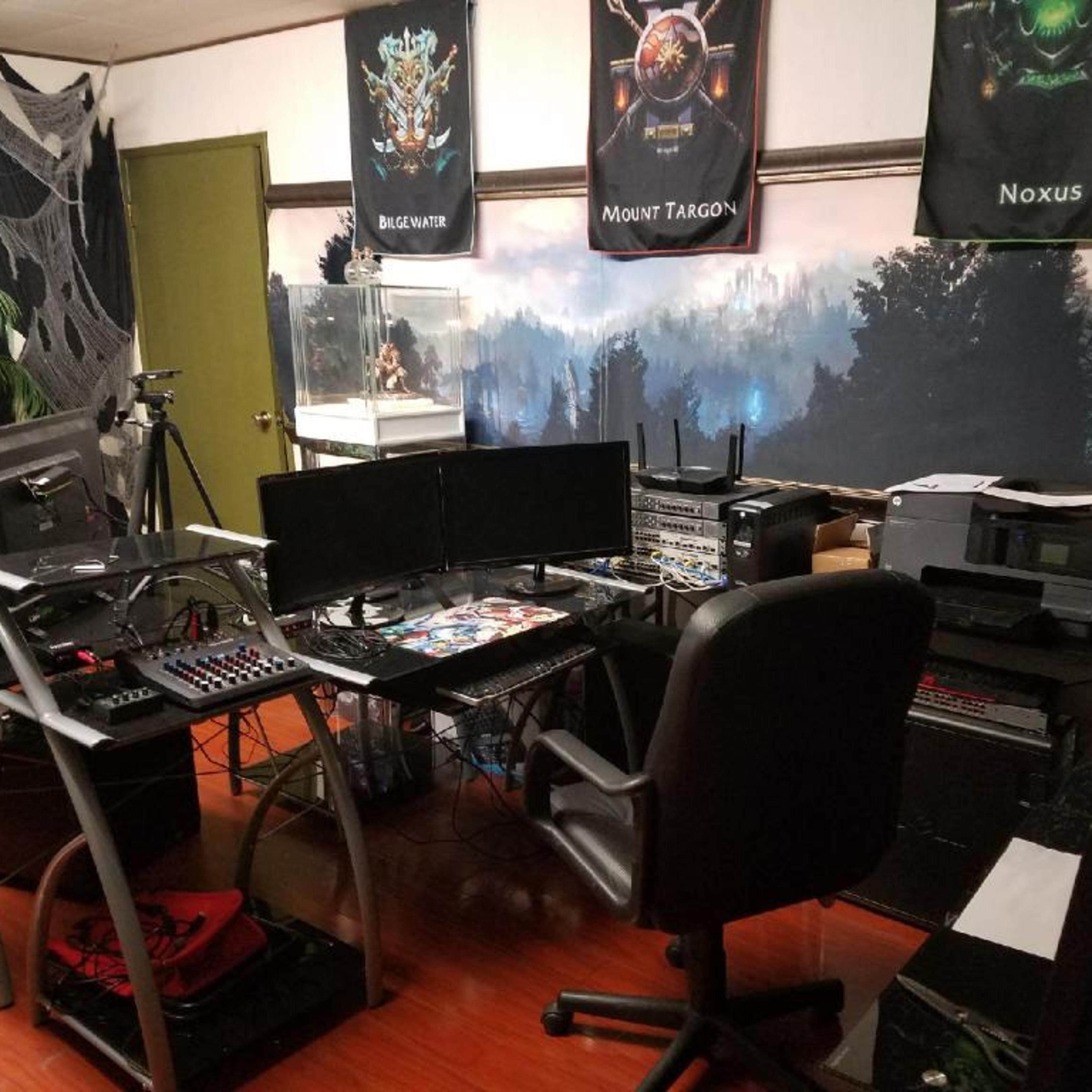 A gaming or tech setup featuring multiple monitors, audio equipment, and a backdrop of a forest scene, with various gaming-related posters on the walls.