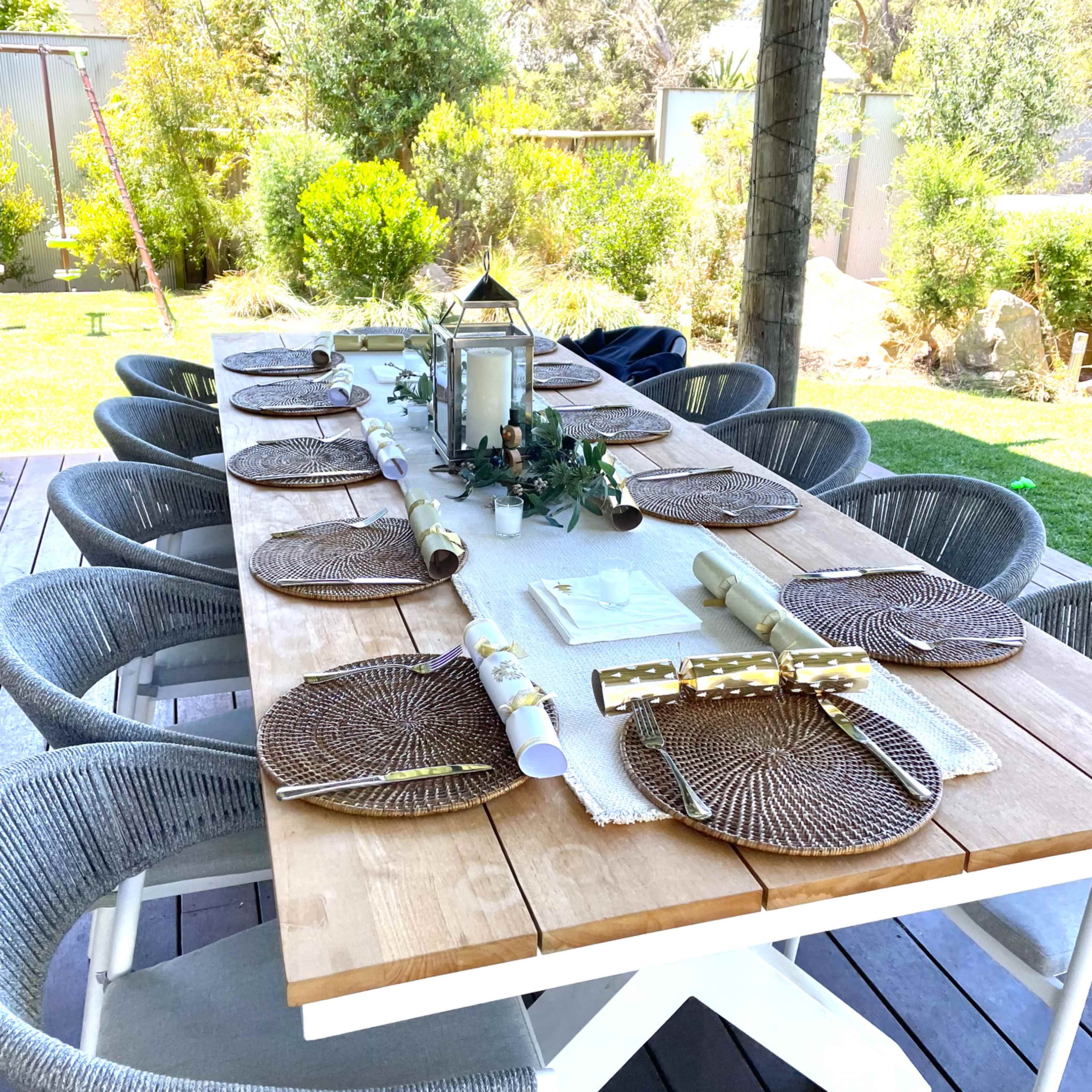 A long wooden dining table is set outdoors with round placemats and decorative items under a shaded area surrounded by greenery.