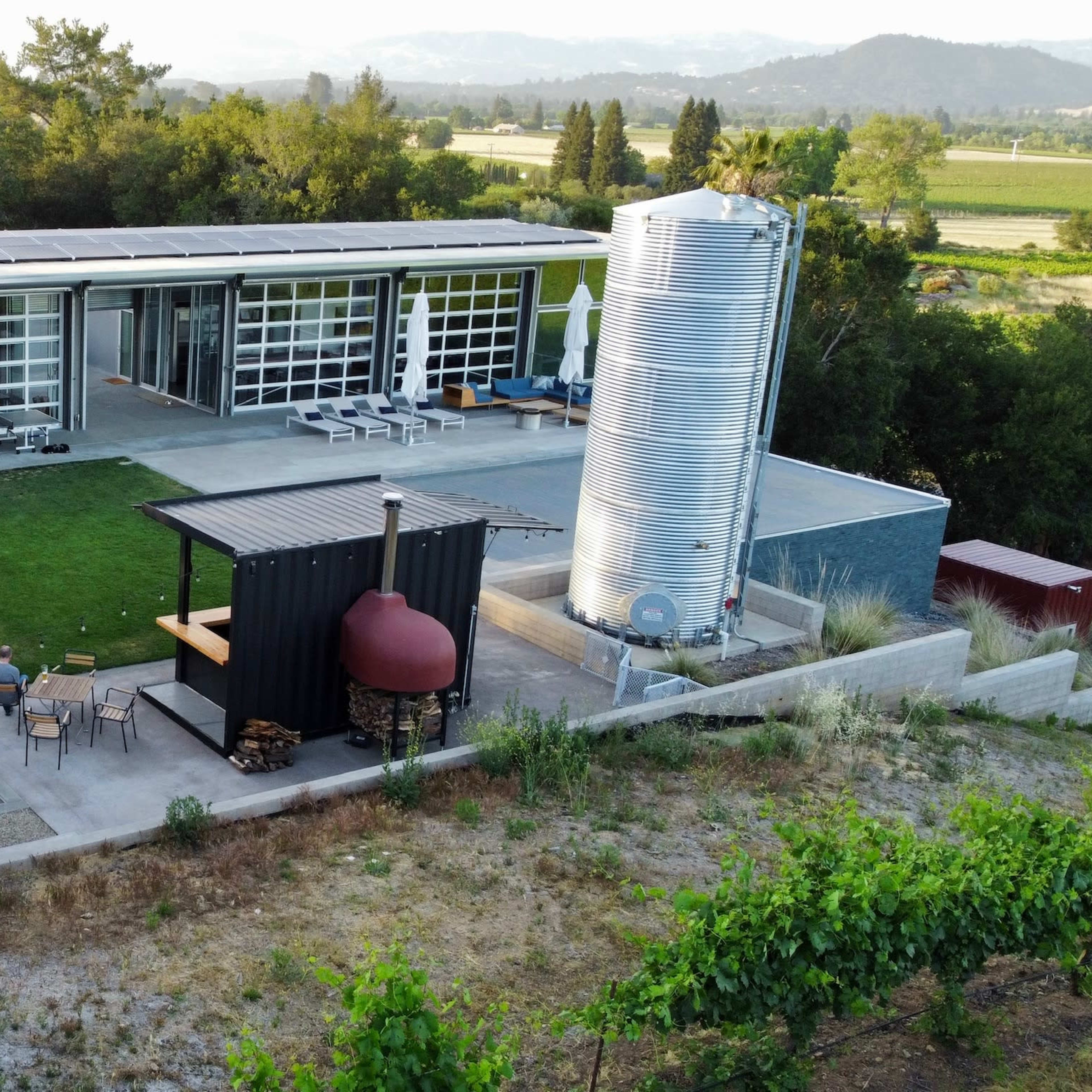 A modern building with large glass windows and solar panels is surrounded by a landscaped area, featuring a seating space and a nearby metal water tank.