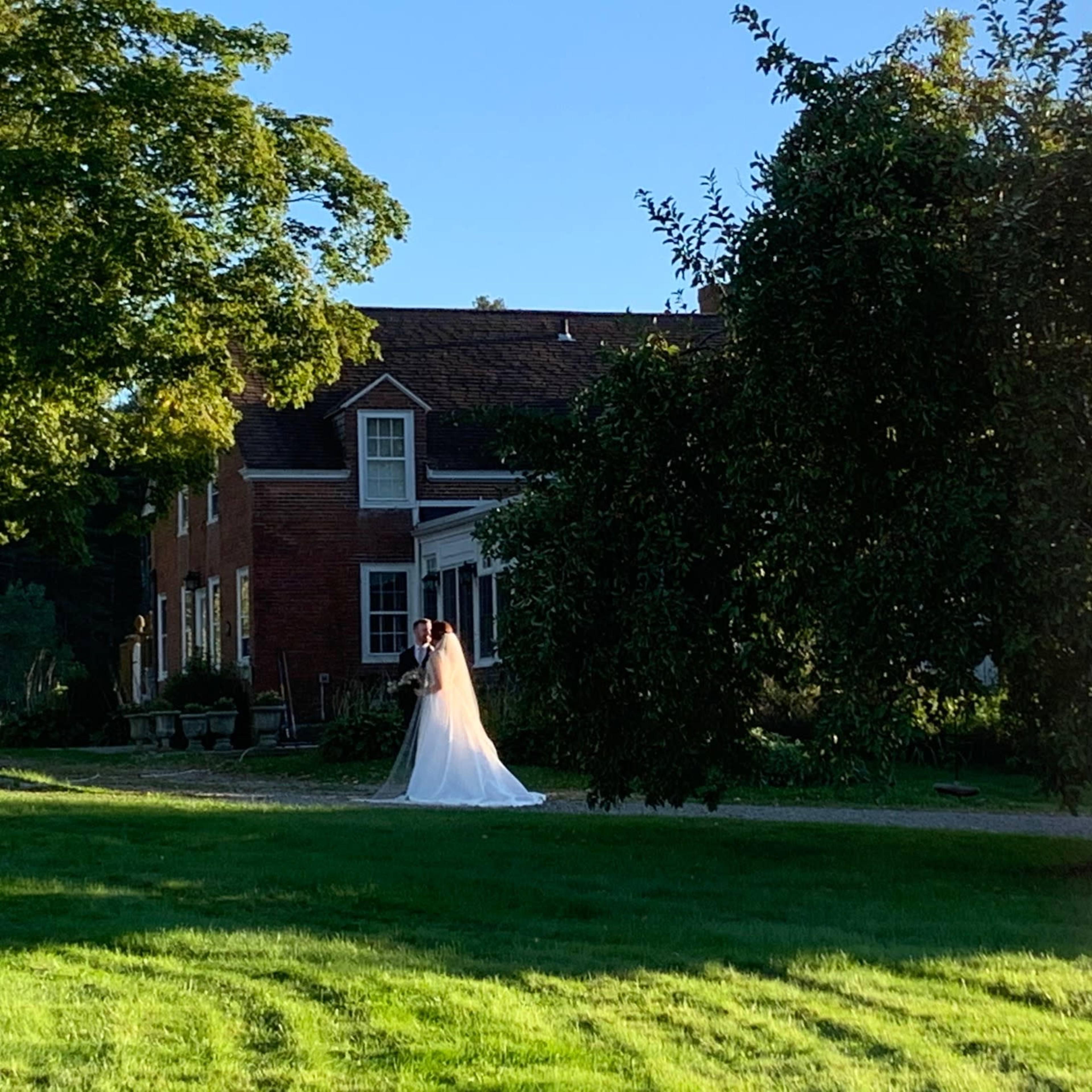 A bride in a white dress stands in a green lawn in front of a brick house.