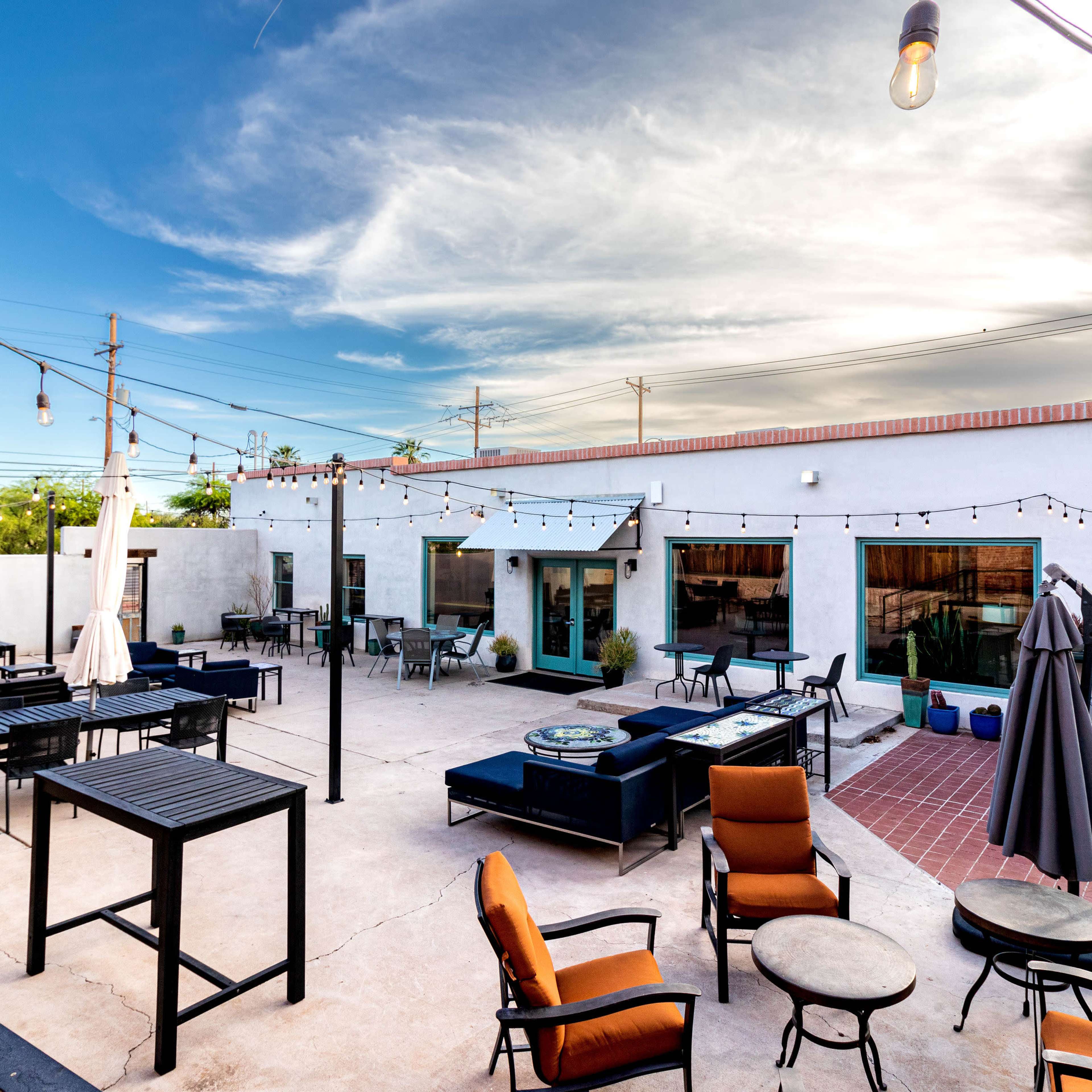 An outdoor patio area with tables and chairs, string lights, and several umbrellas under a clear blue sky.
