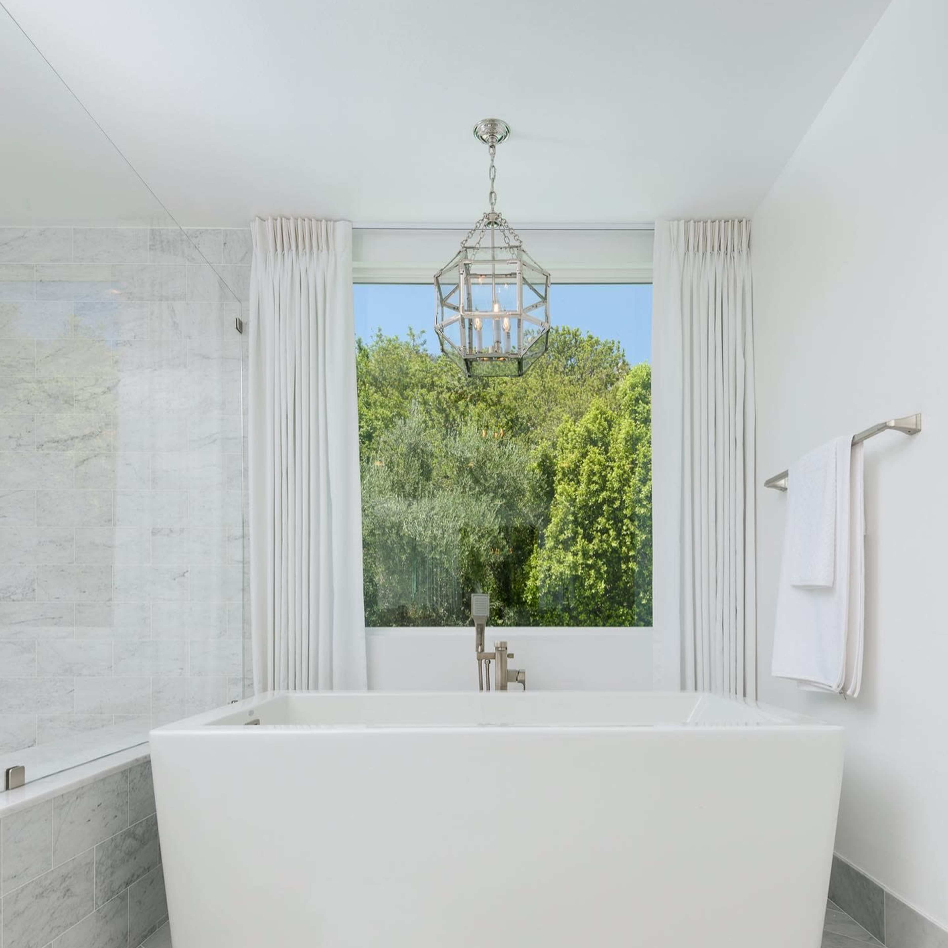 A modern bathroom features a freestanding white bathtub, large windows overlooking greenery, and marble tile flooring.