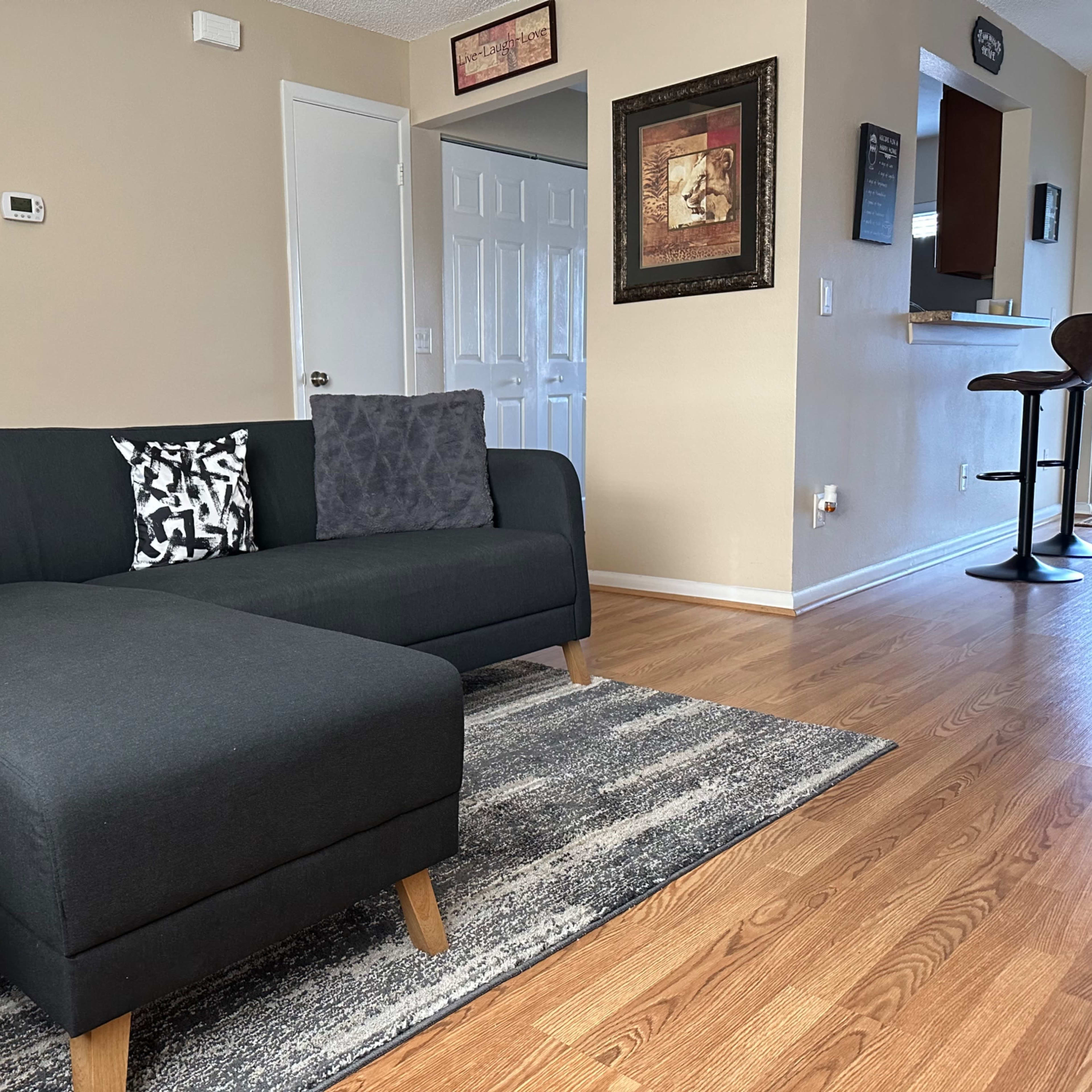 A modern living room featuring a black sectional sofa with decorative pillows, a patterned rug, and a bar area in the background.