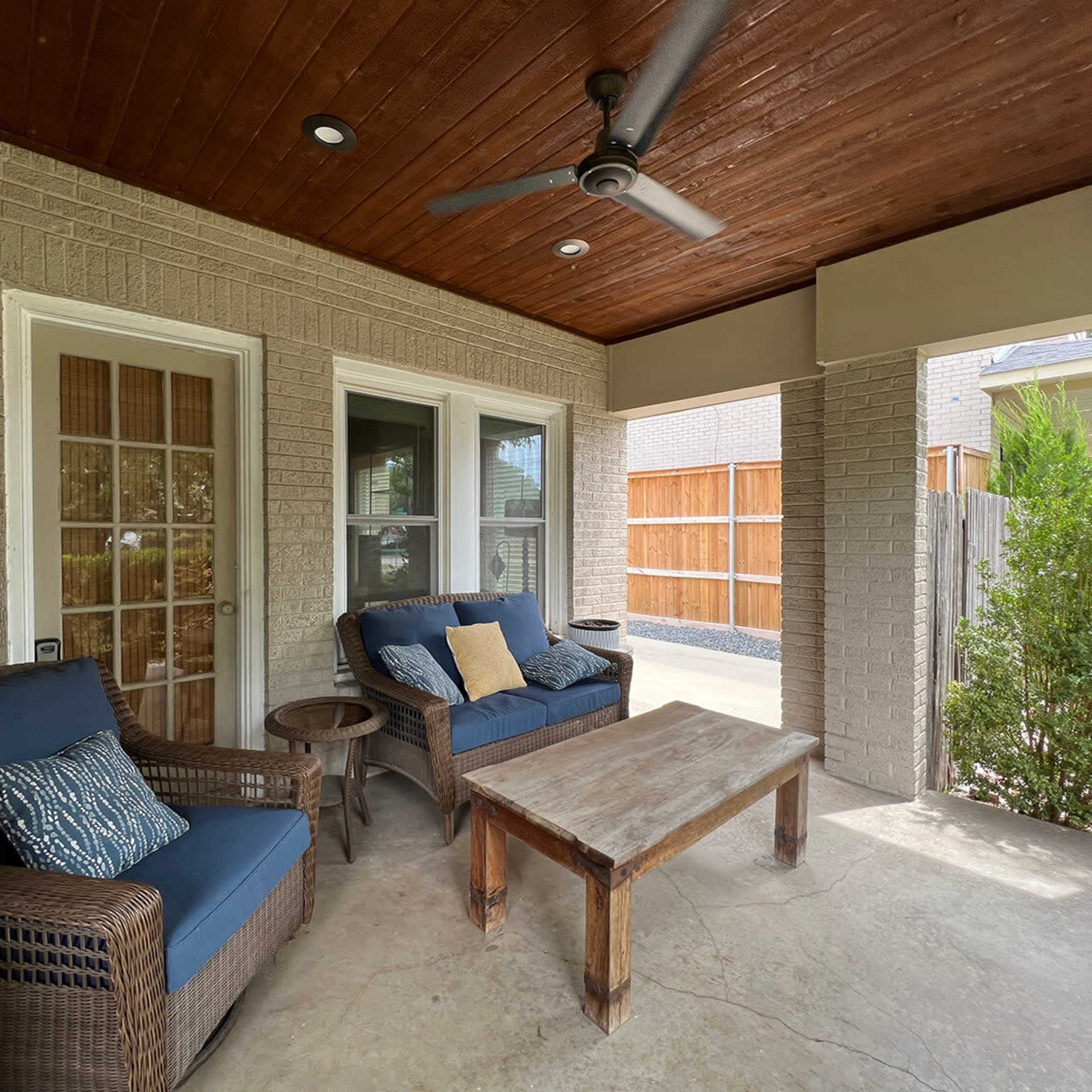 A covered patio area with two blue cushioned chairs, a wooden coffee table, and a ceiling fan.