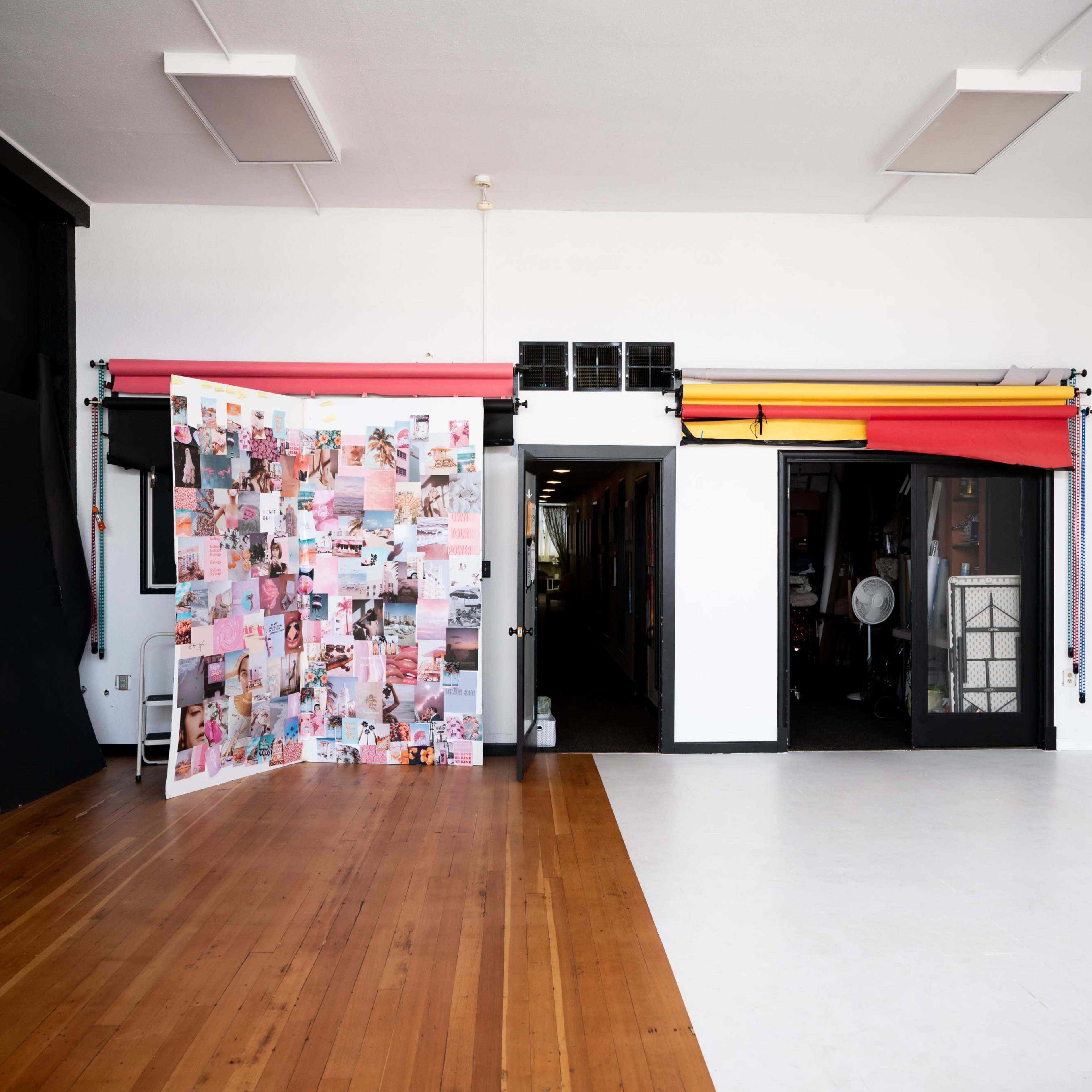 A spacious studio with a wooden floor, featuring a collage panel on the left and a brightly lit entrance to another room adorned with colorful backdrops.